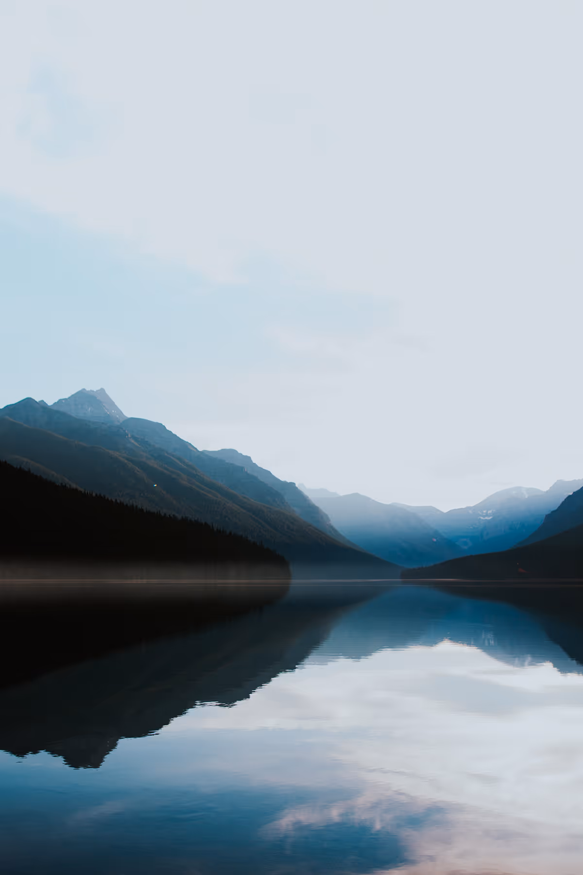 Calm lake reflecting mountain ranges and a pale sky with clouds.