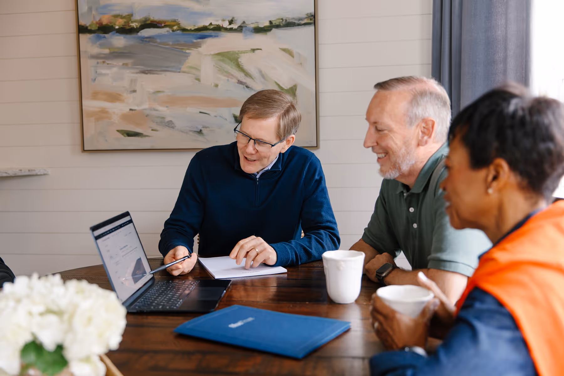 Three adults sitting around a table, looking at a laptop screen during a discussion, with coffee mugs and a folder on the table.