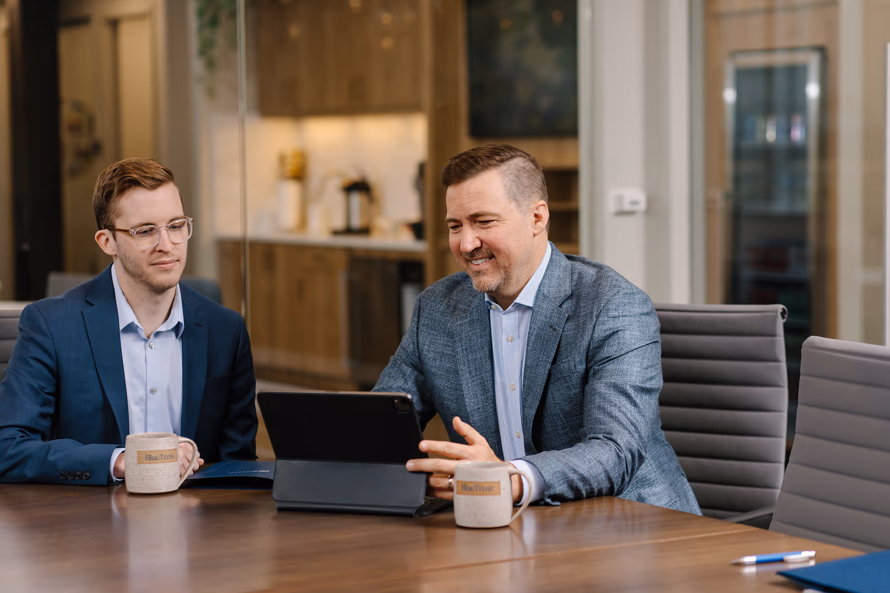 Two men in business attire sitting at a conference table with mugs, looking at a tablet together in an office setting.