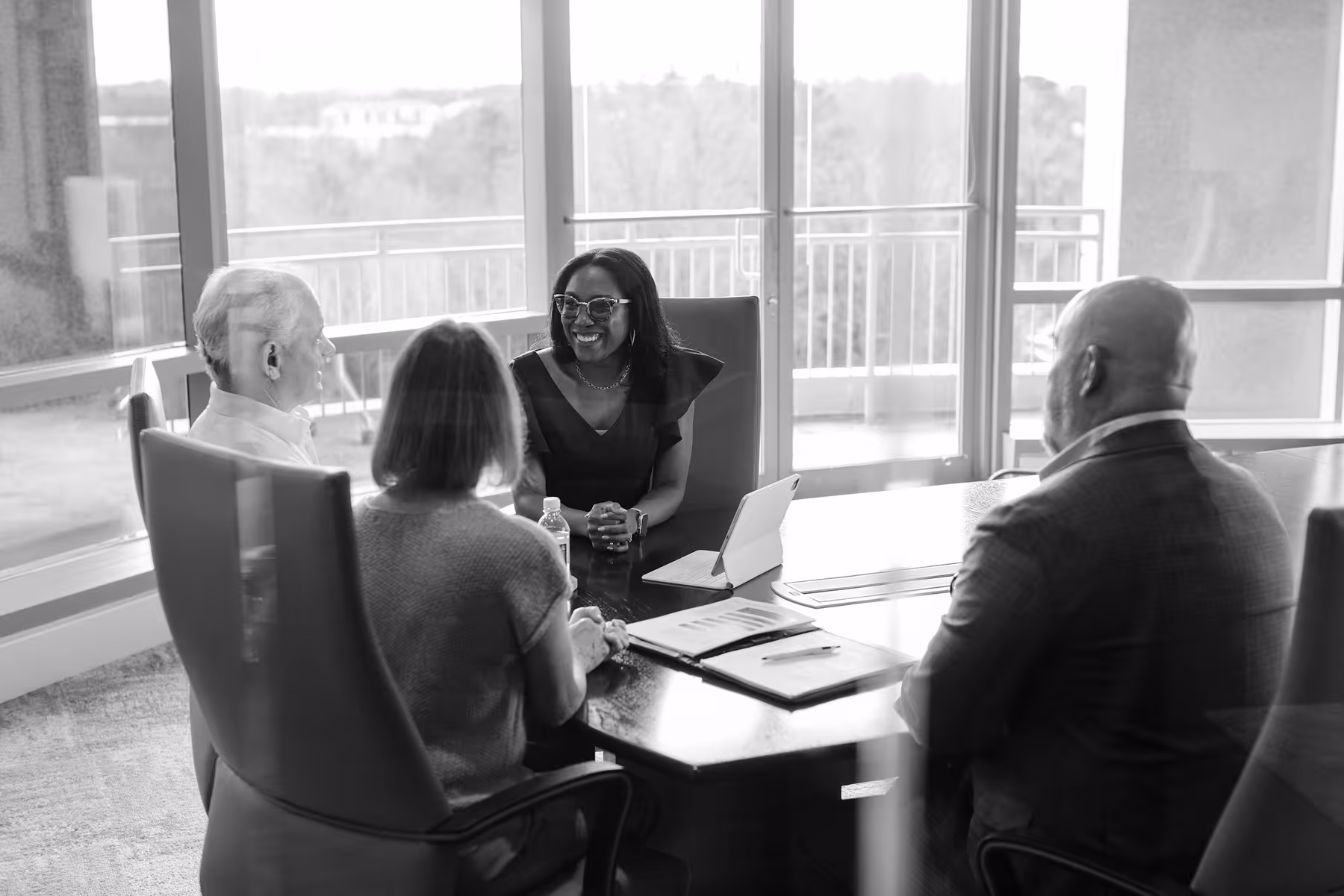 Four diverse professionals engaged in a meeting around a conference table with documents and a tablet in a modern office with large windows.