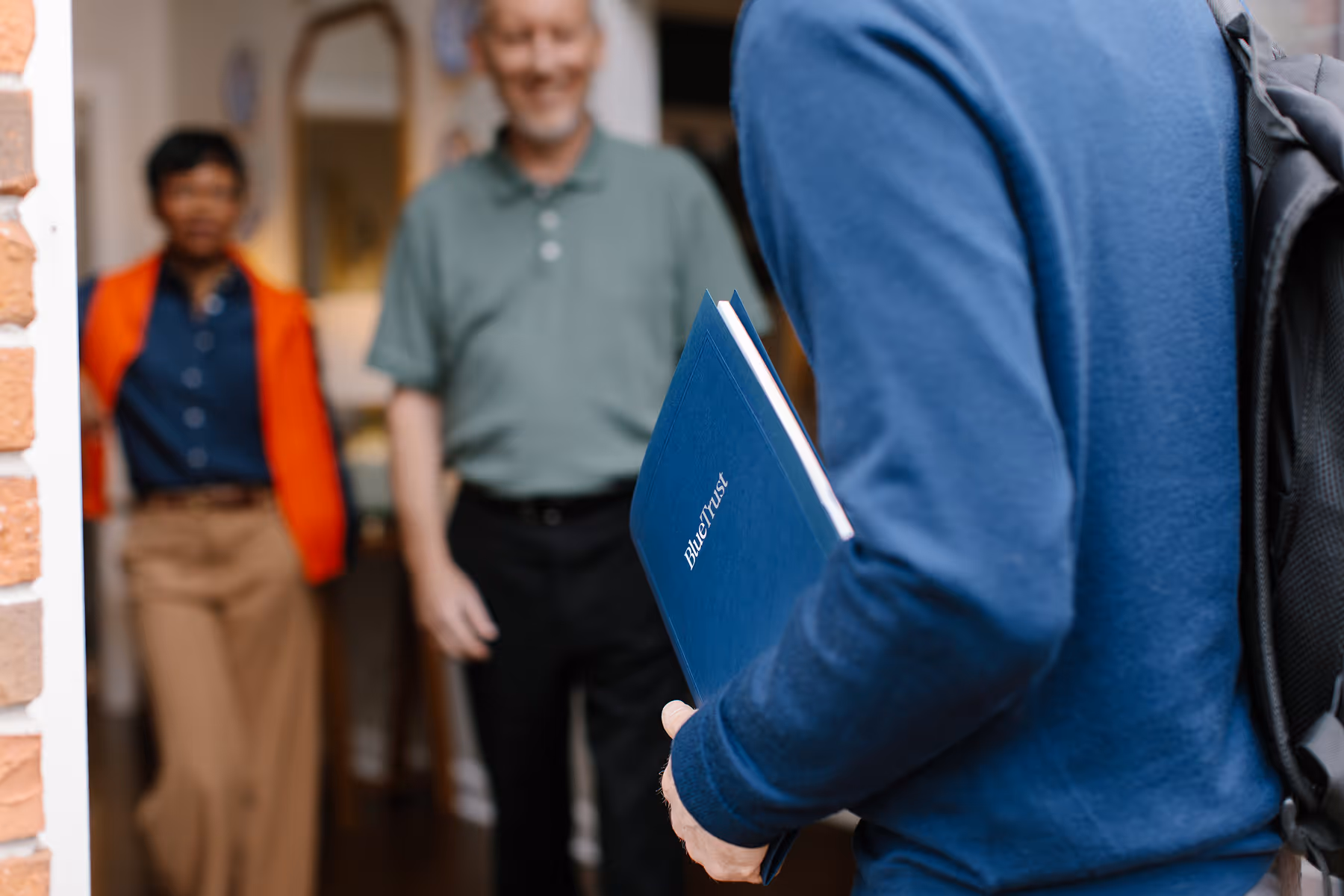 Person in blue holding a BlueTrust folder standing in a doorway with two people smiling inside a house.