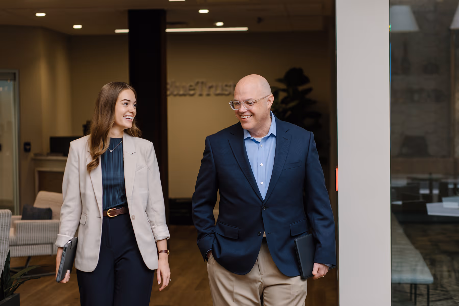 Two business colleagues smiling and walking inside an office, each holding a closed laptop.
