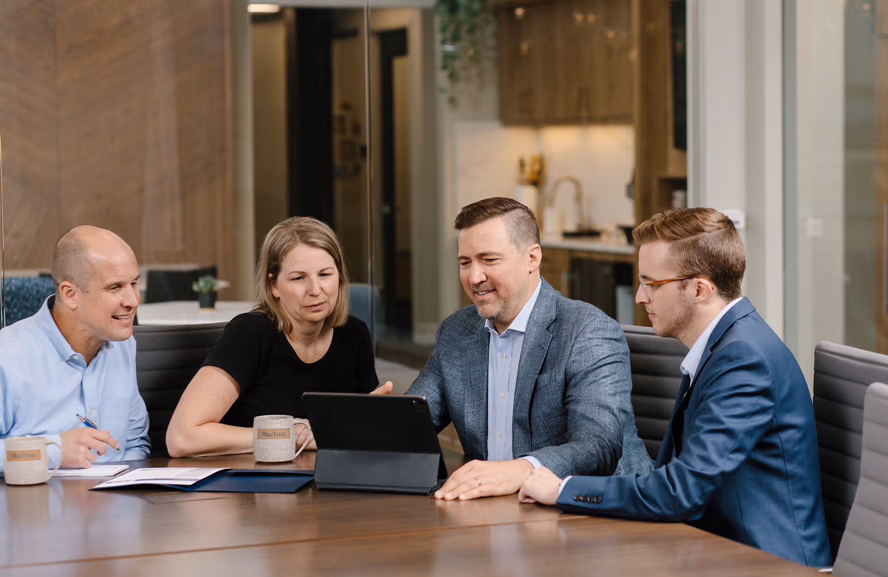Four professionals in a modern office discussing work around a conference table with a tablet and documents.