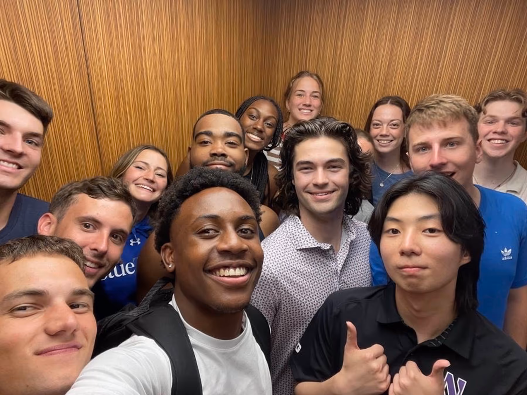 Group of smiling young adults posing closely for a selfie in an elevator with wood panel walls.