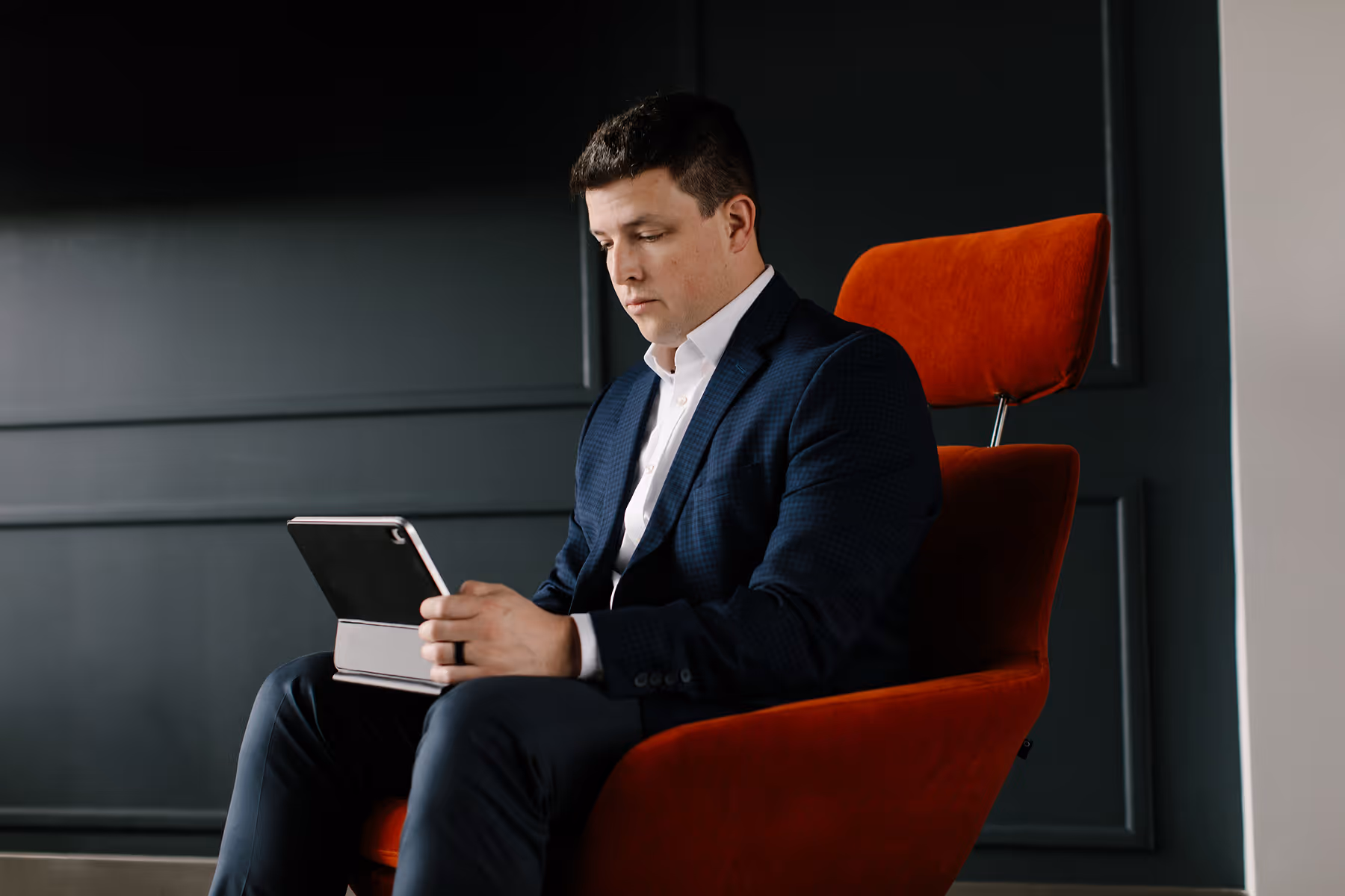 Man in navy suit sitting in orange chair, focused on tablet in his hands.