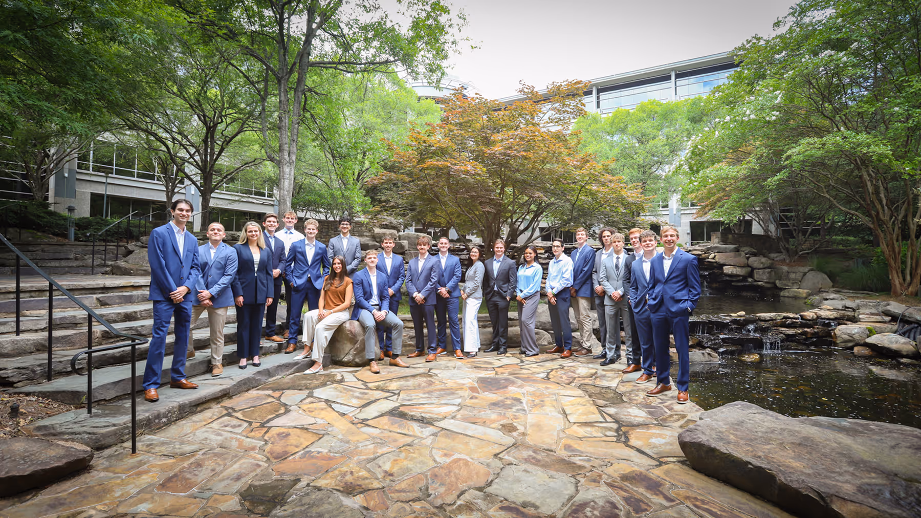 Group of young professionals and interns in business attire posing outdoors near pond and trees.