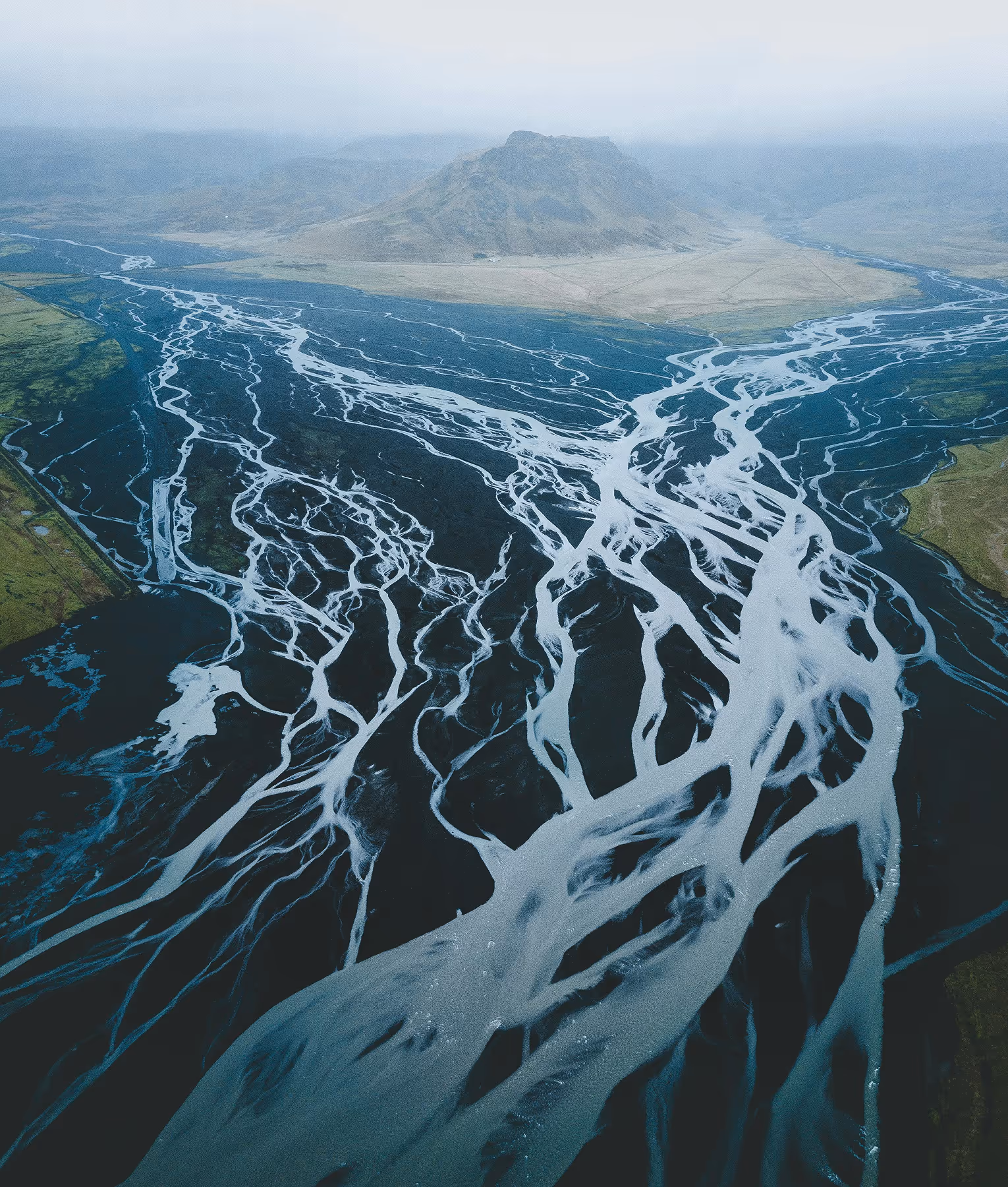 A braided river with multiple channels flowing through a dark landscape with a mountain in the background.