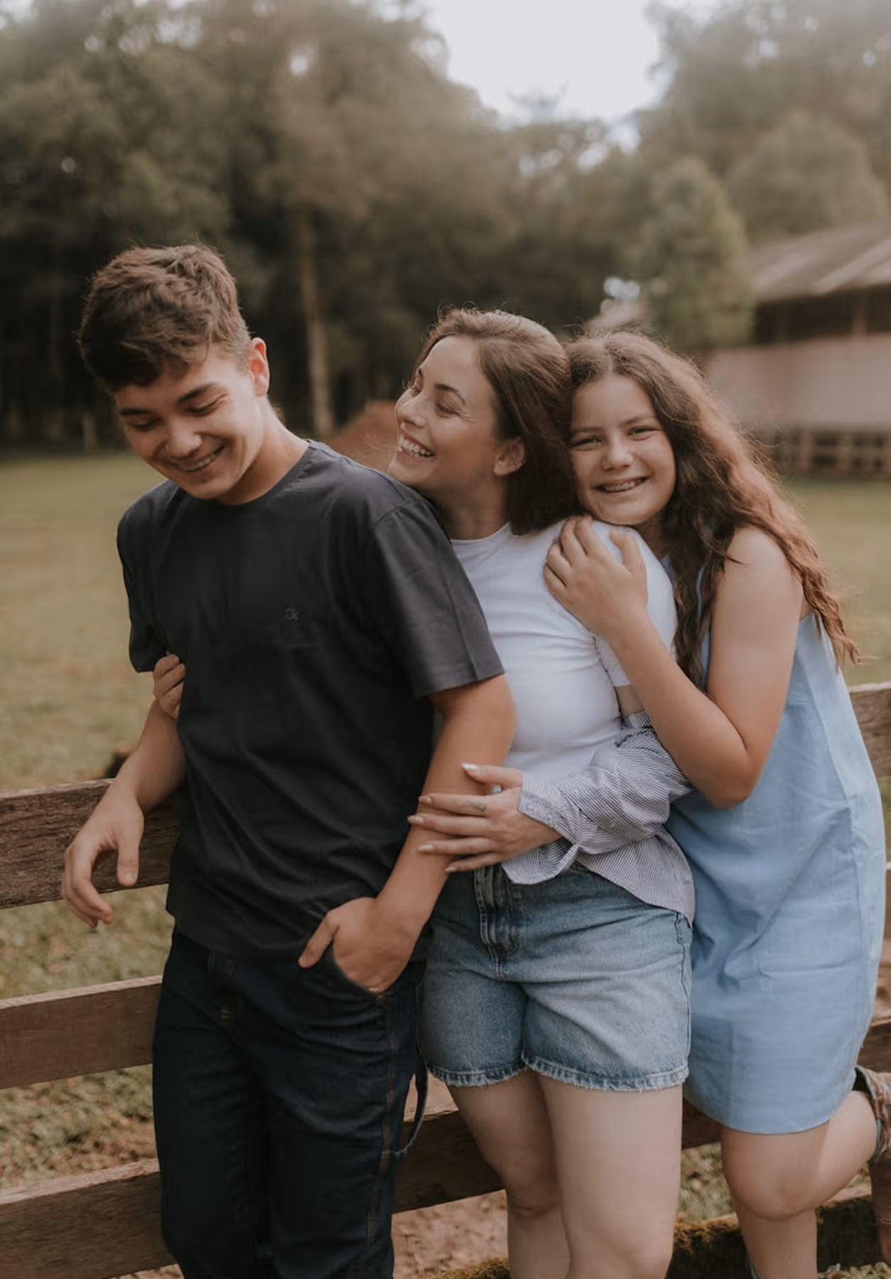 Three smiling kids standing outdoors near a wooden fence with trees in the background.