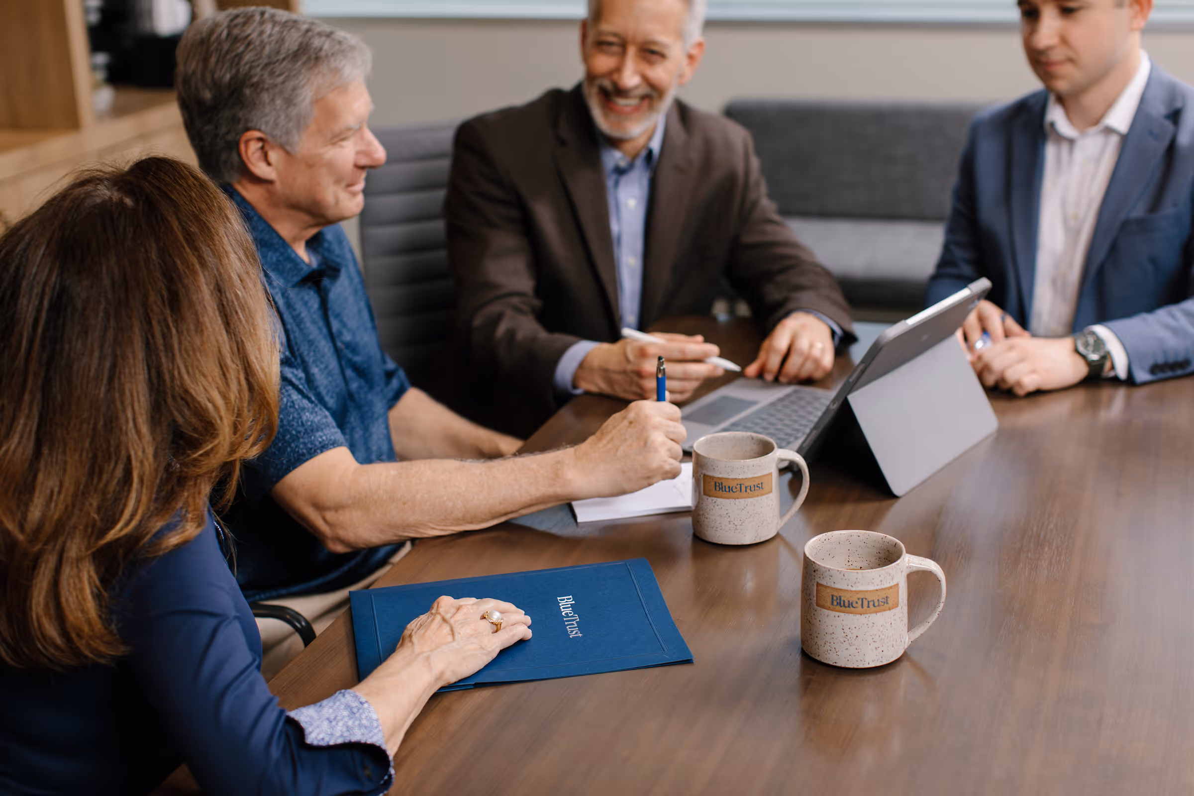 Four people in a meeting at a table with BlueTrust folders, mugs, and a tablet with keyboard.
