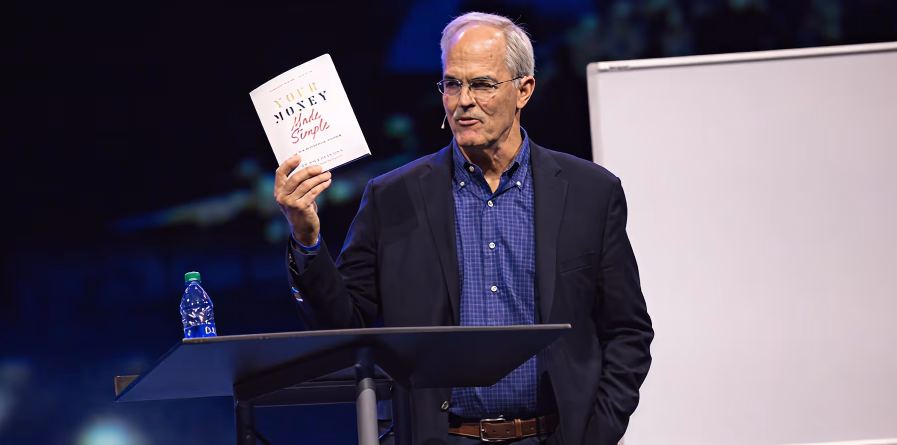 Man in glasses and blazer holds up a book titled Your Money Made Simple while speaking on stage.