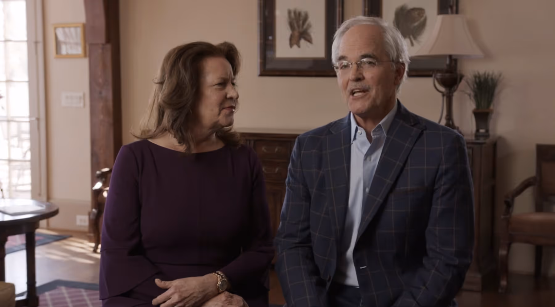 Older couple sitting indoors, the woman looks at the man who is speaking, both dressed formally.