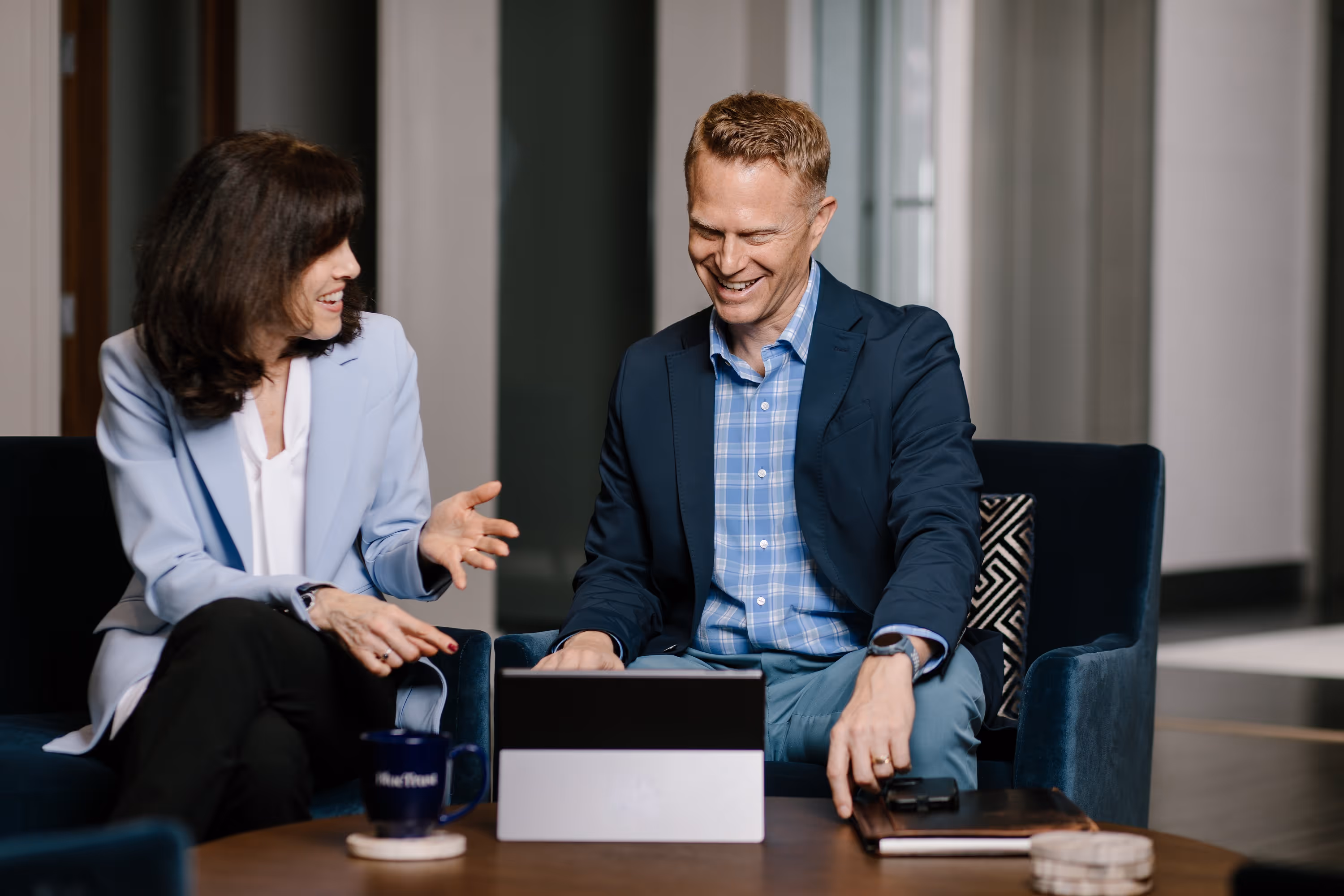 Two businesspeople smiling and talking while using a laptop in a modern office lounge.