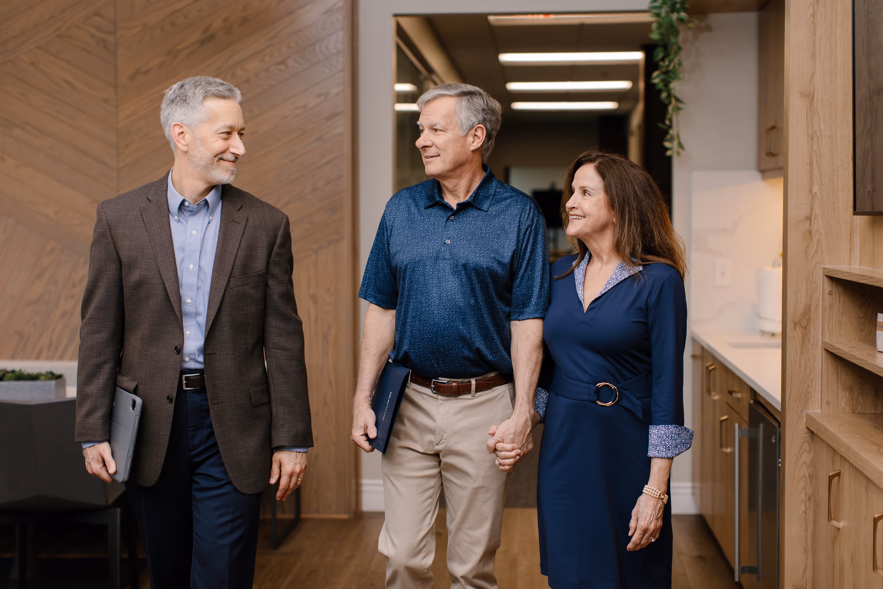 Man in blazer walking with smiling couple holding hands down a hallway.