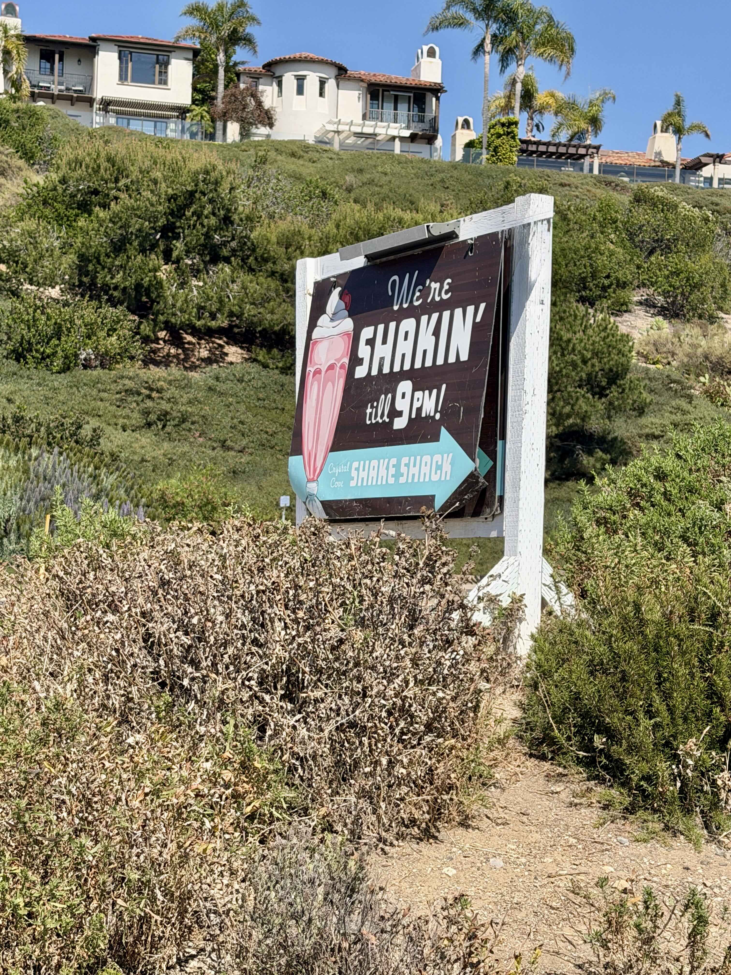 Vintage 'We're Shakin' till 9PM' wooden sign along Pacific Coast Highway pointing to Crystal Cove Shake Shack, with Crystal Cove hillside homes in the background