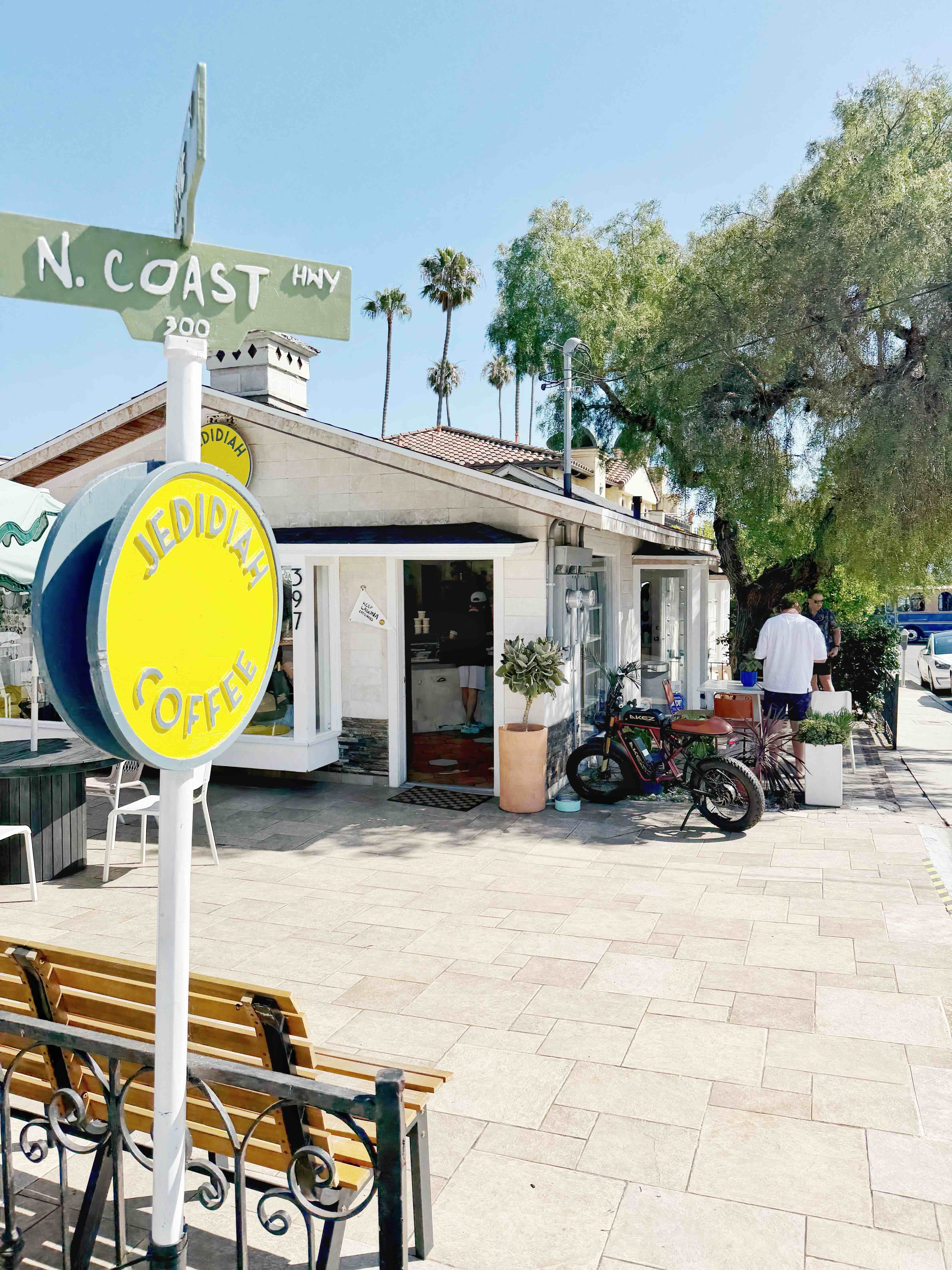 Jedidiah Coffee at 397 N Coast Highway, Laguna Beach — yellow circle sign at the curb beside the N. Coast Hwy street sign, with wooden bench and parked e-bikes outside