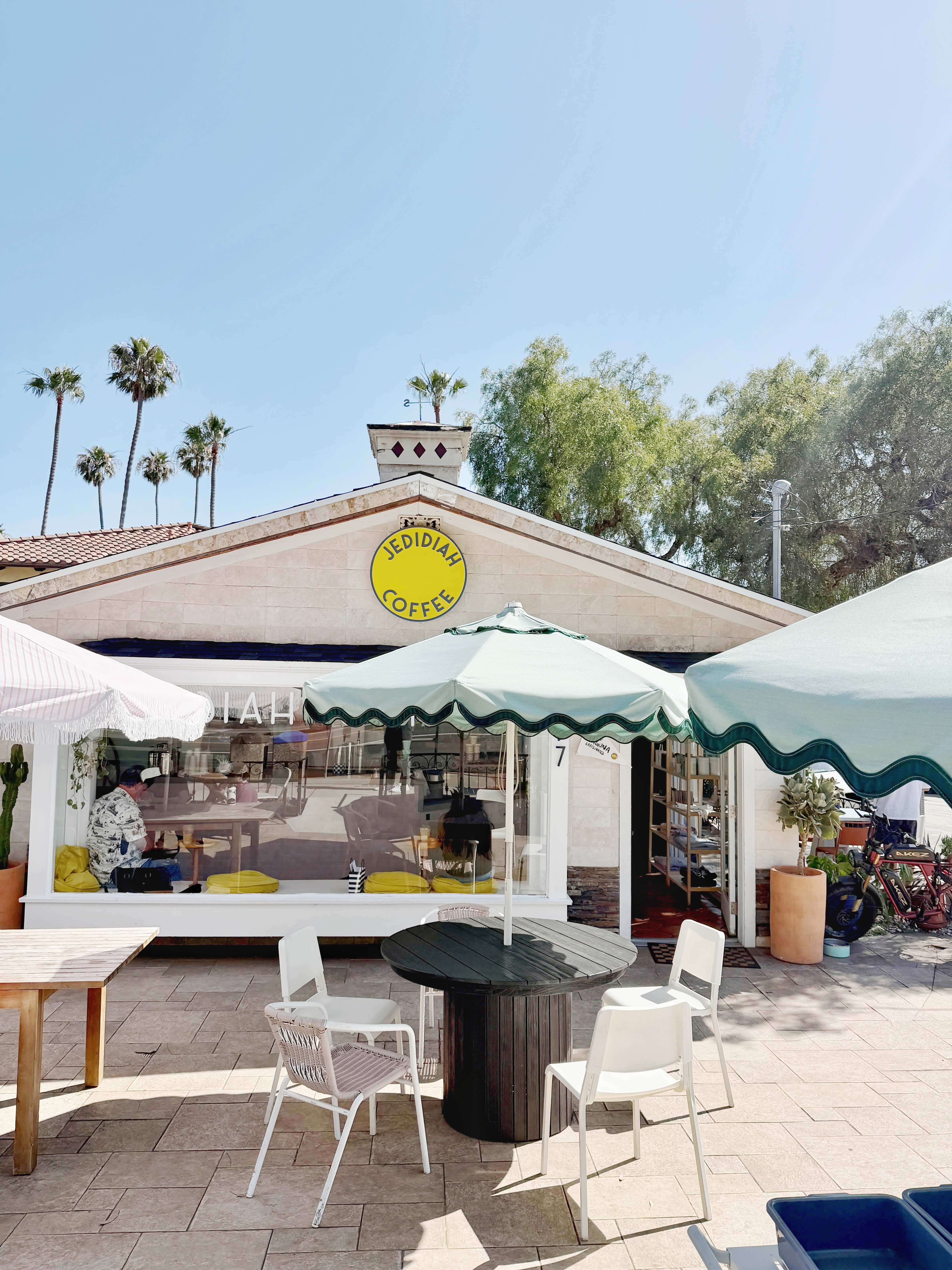 Jedidiah Coffee storefront in downtown Laguna Beach — white craftsman building with yellow Jedidiah Coffee circle logo sign, scalloped sage umbrellas, and patio seating on a sunny day