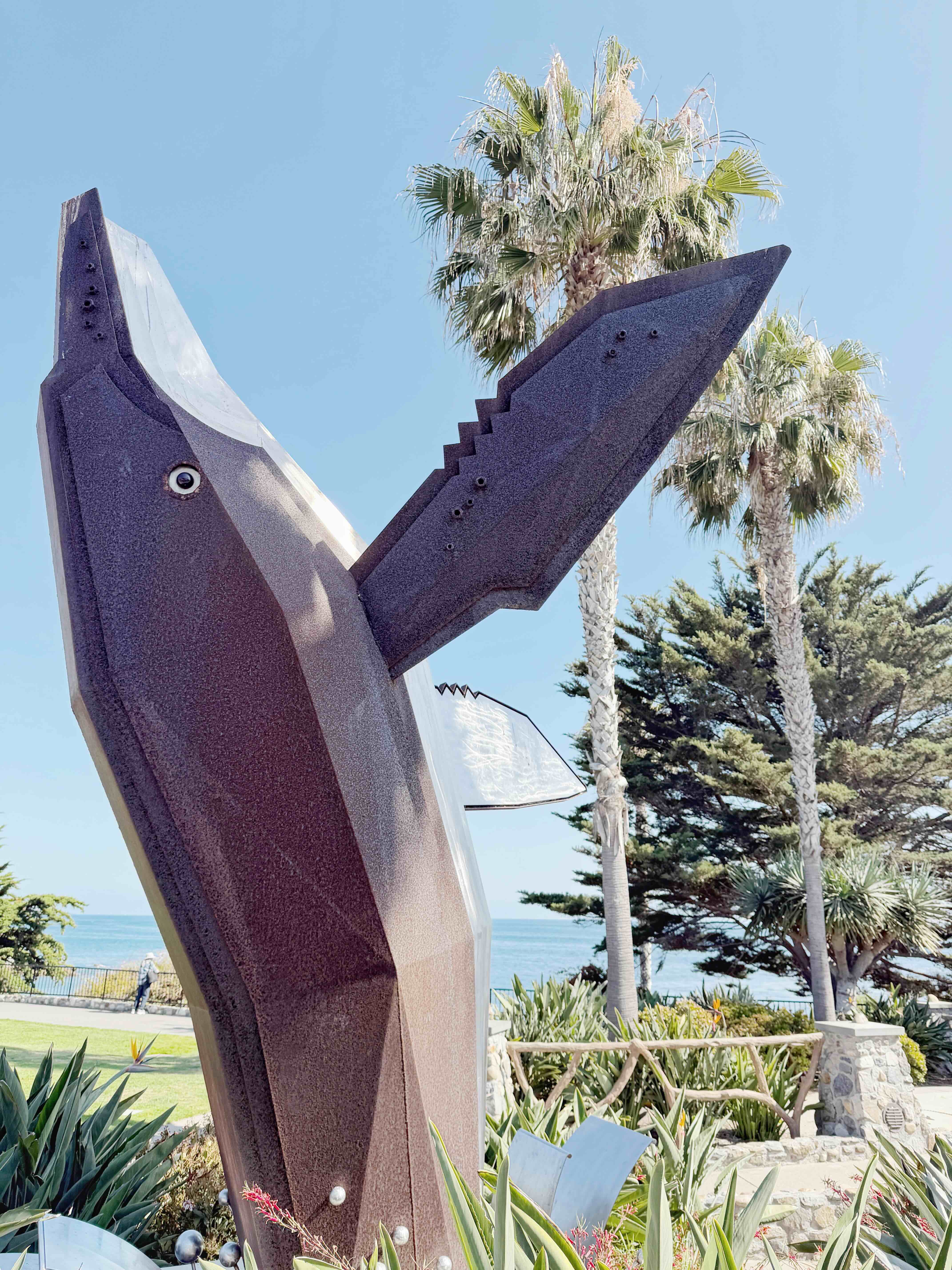 Heisler Park's iconic metal whale sculpture against blue sky and palm trees, with a glimpse of the Pacific Ocean in the background