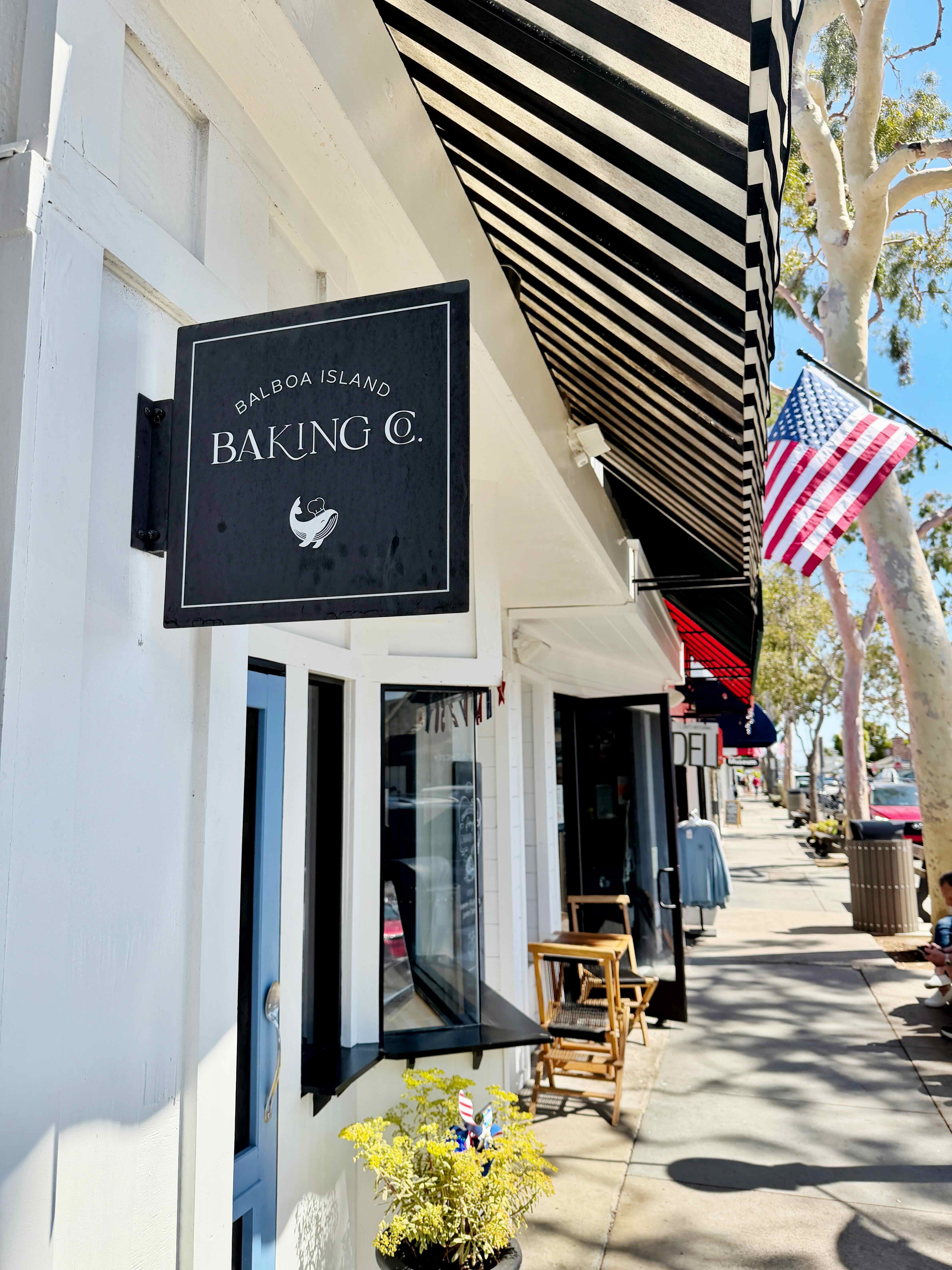 Balboa Island Baking Co. hanging black sign with the bird logo, mounted under a black and white striped awning on Marine Avenue, American flag visible above