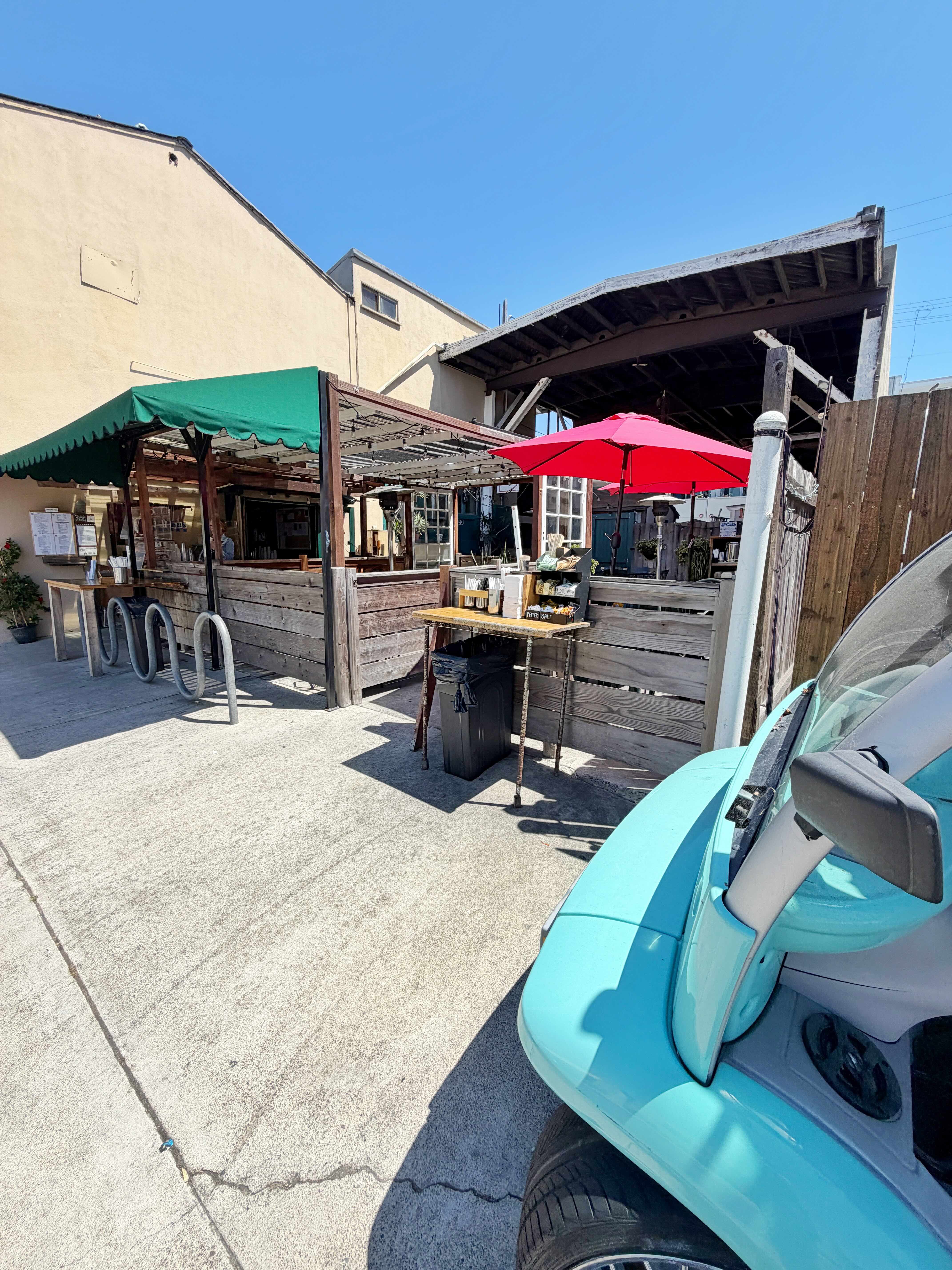 Alta Coffee back patio in Newport Beach — green awning, wooden benches, a red umbrella over a tasting counter, and a teal Polaris golf cart parked nearby