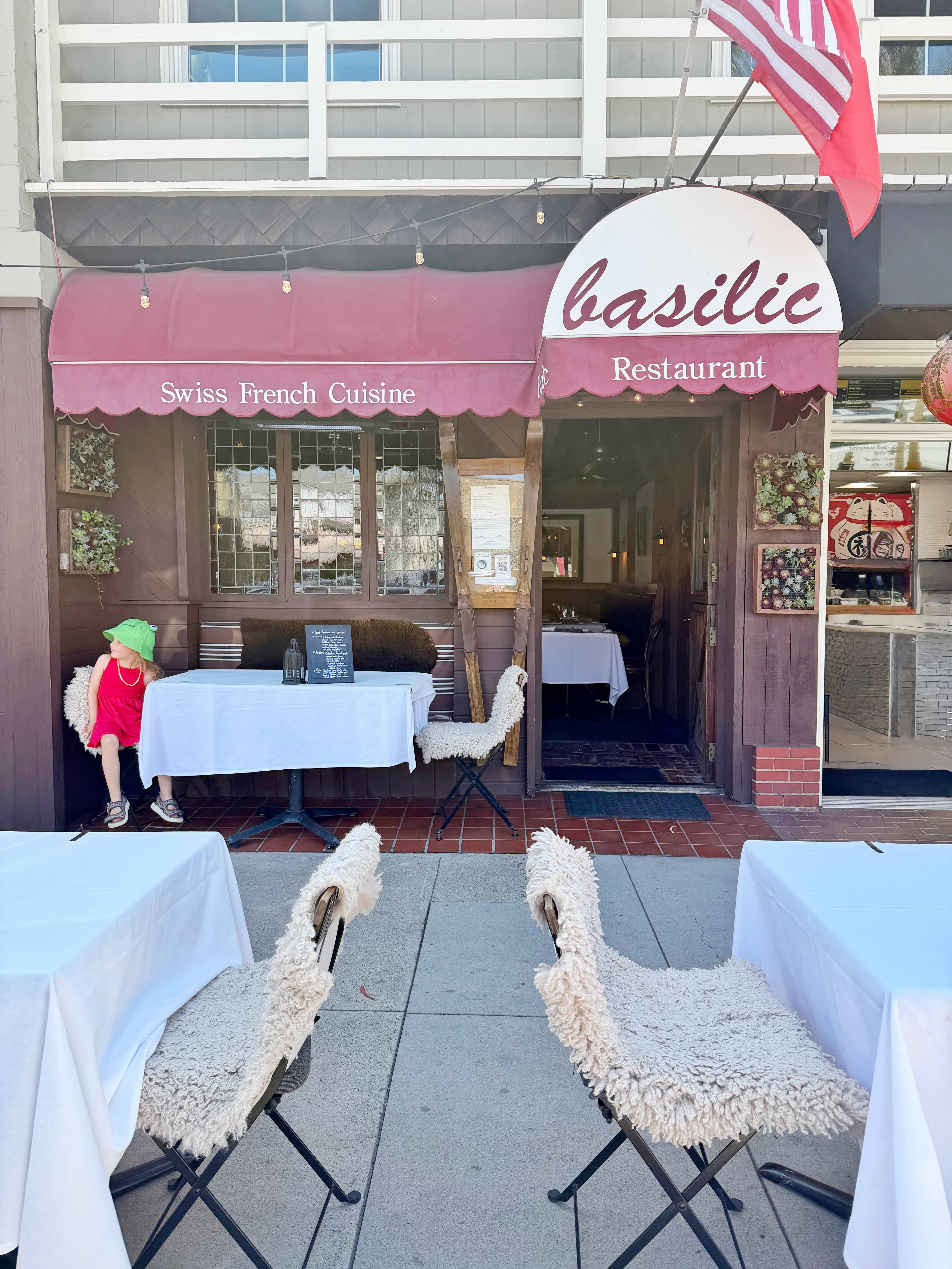 Basilic Restaurant on Balboa Island — burgundy awning over the Swiss-French dining storefront on Marine Avenue with sidewalk tables, fluffy white chair covers, and the American flag flying above