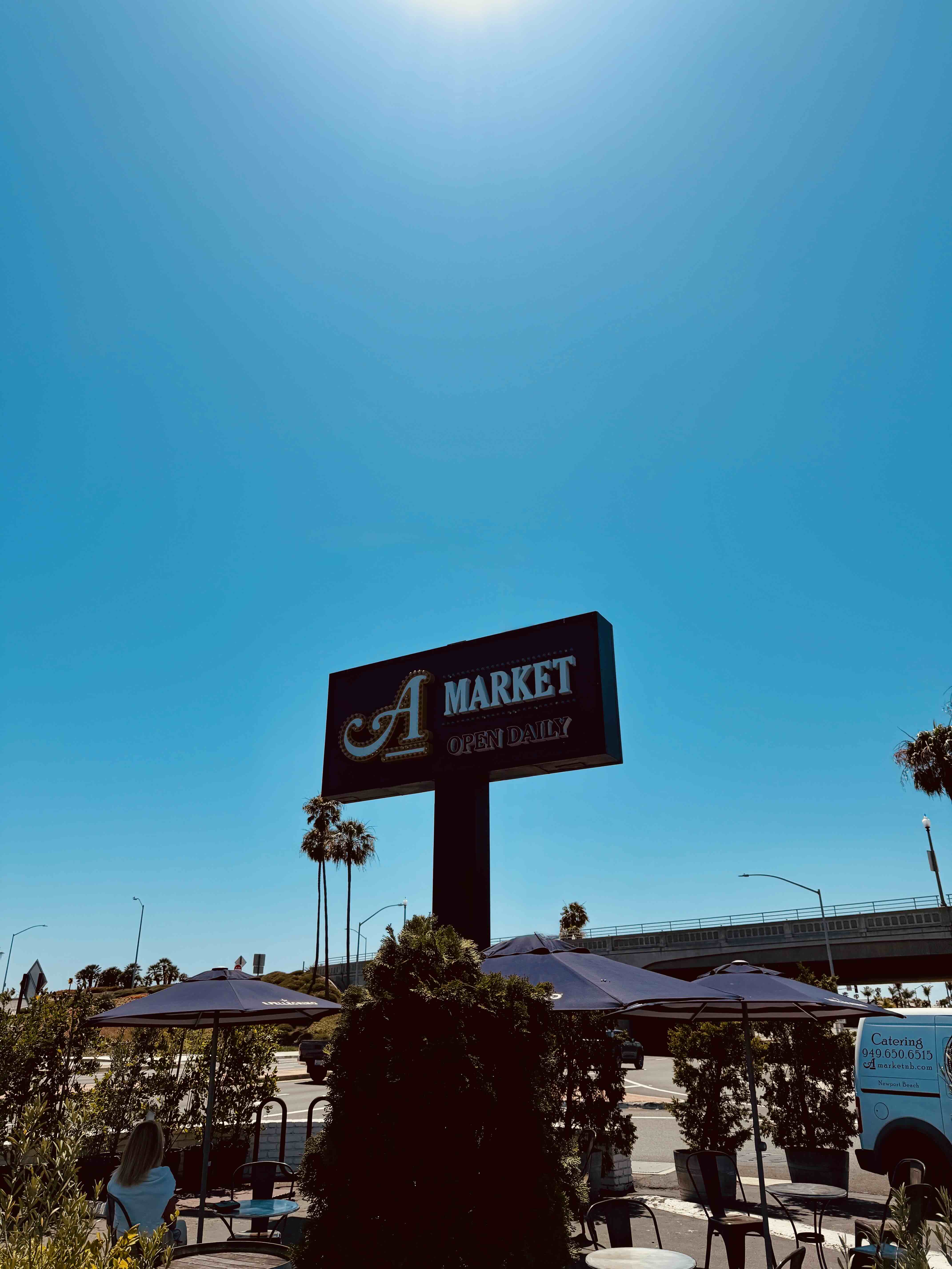 A Market signage along West Coast Highway in Newport Beach — black sign with white 'A Market Open Daily' lettering against a sunny blue sky, with patio umbrellas and palm trees visible below