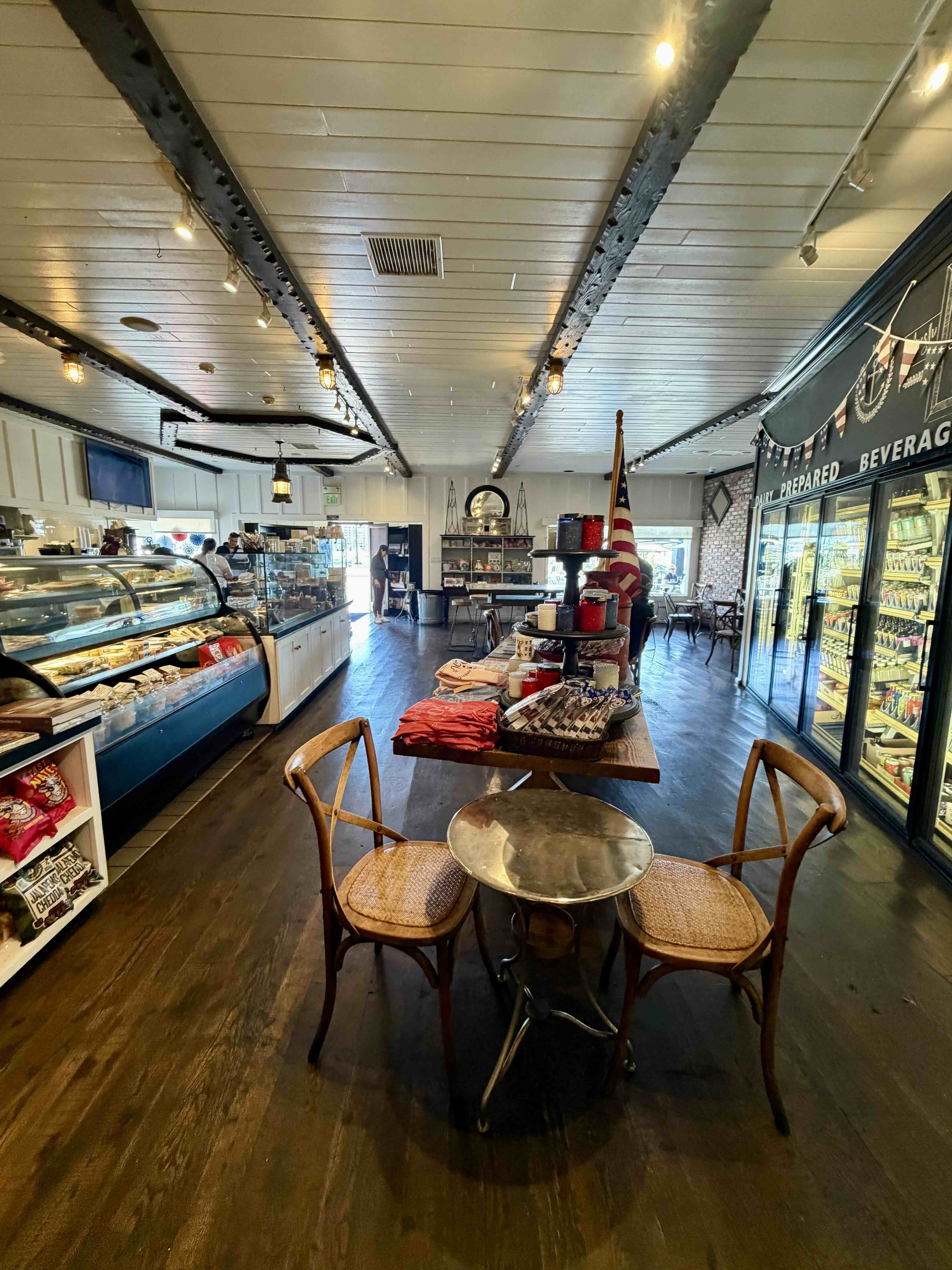 A Market interior in Newport Beach — wood floors and exposed beam ceiling, deli cases on the left, beverage cooler labeled Prepared Beverages on the right, with small bistro seating in the foreground