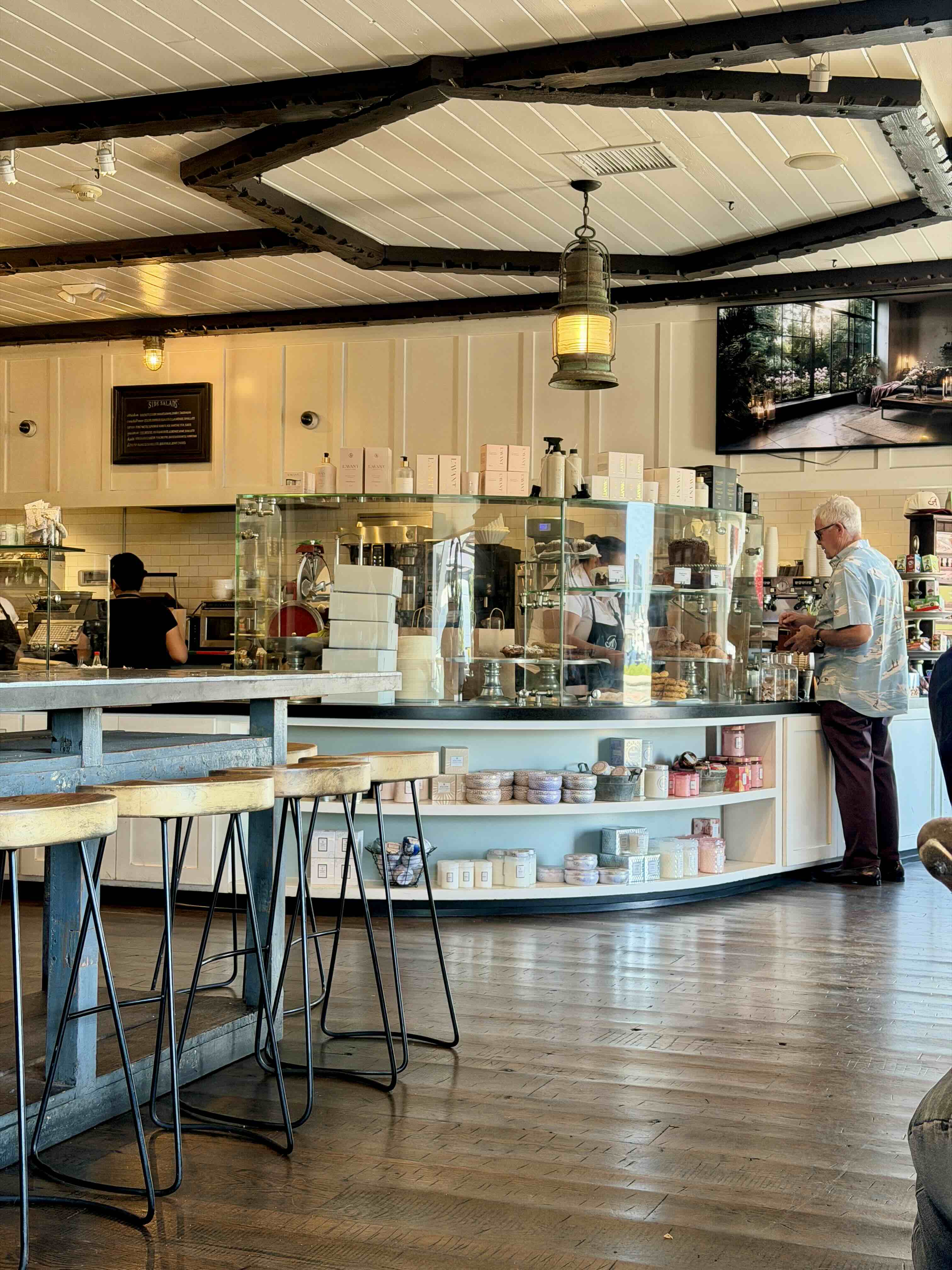 A Market central counter and bakery case in Newport Beach — coastal-painted island base under exposed beams with pendant lighting, counter stools, and a staff member helping a customer