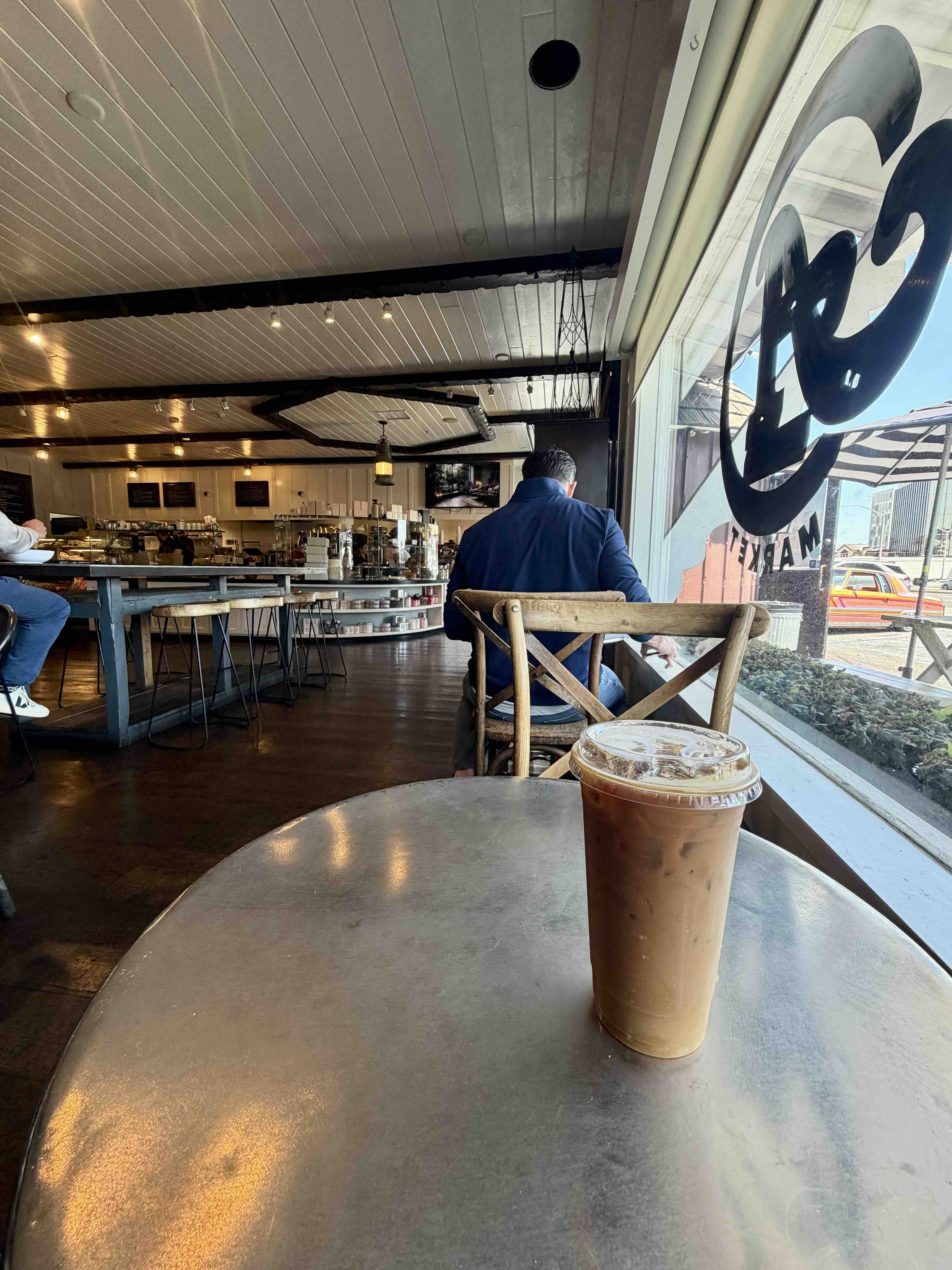 Iced coffee on a small wood table at A Market Newport Beach — view through the window with the A Market logo decal visible and a seated customer nearby