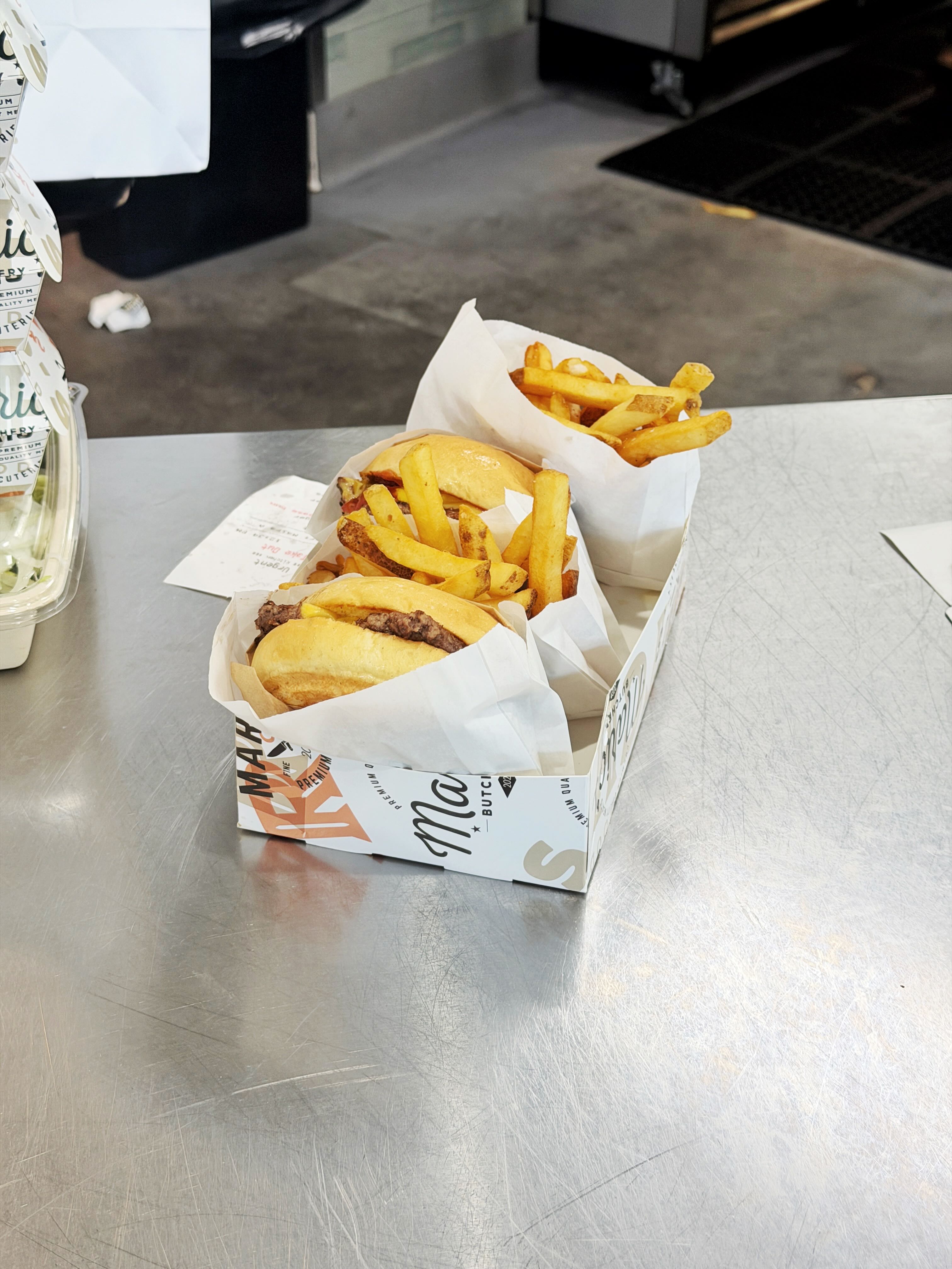 Two Mario's Butcher Shop smash burgers and fries served in a Mario's-branded paper box on a stainless prep counter