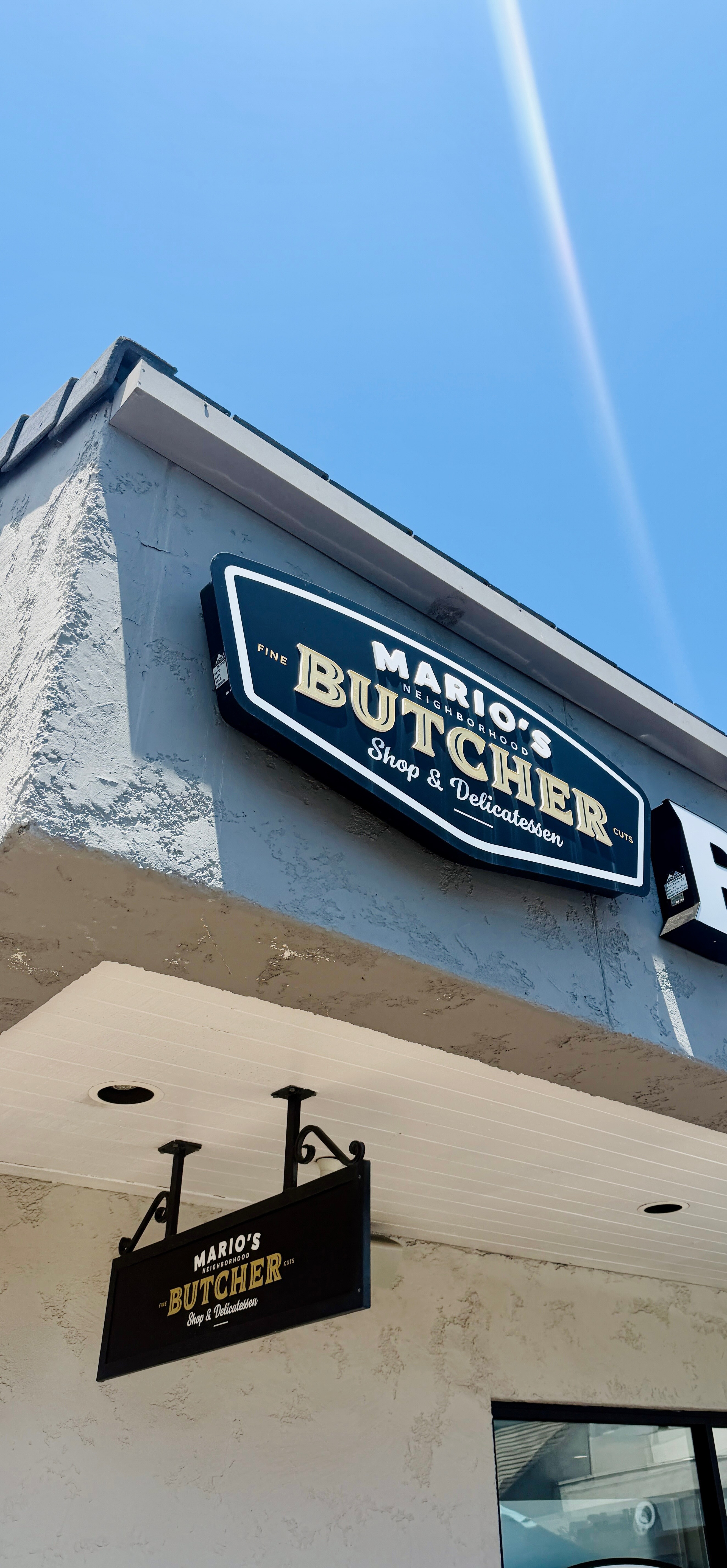 Mario's Neighborhood Butcher Shop & Delicatessen building sign in Newport Beach — navy and cream lettering on the storefront corner with a smaller hanging sign below