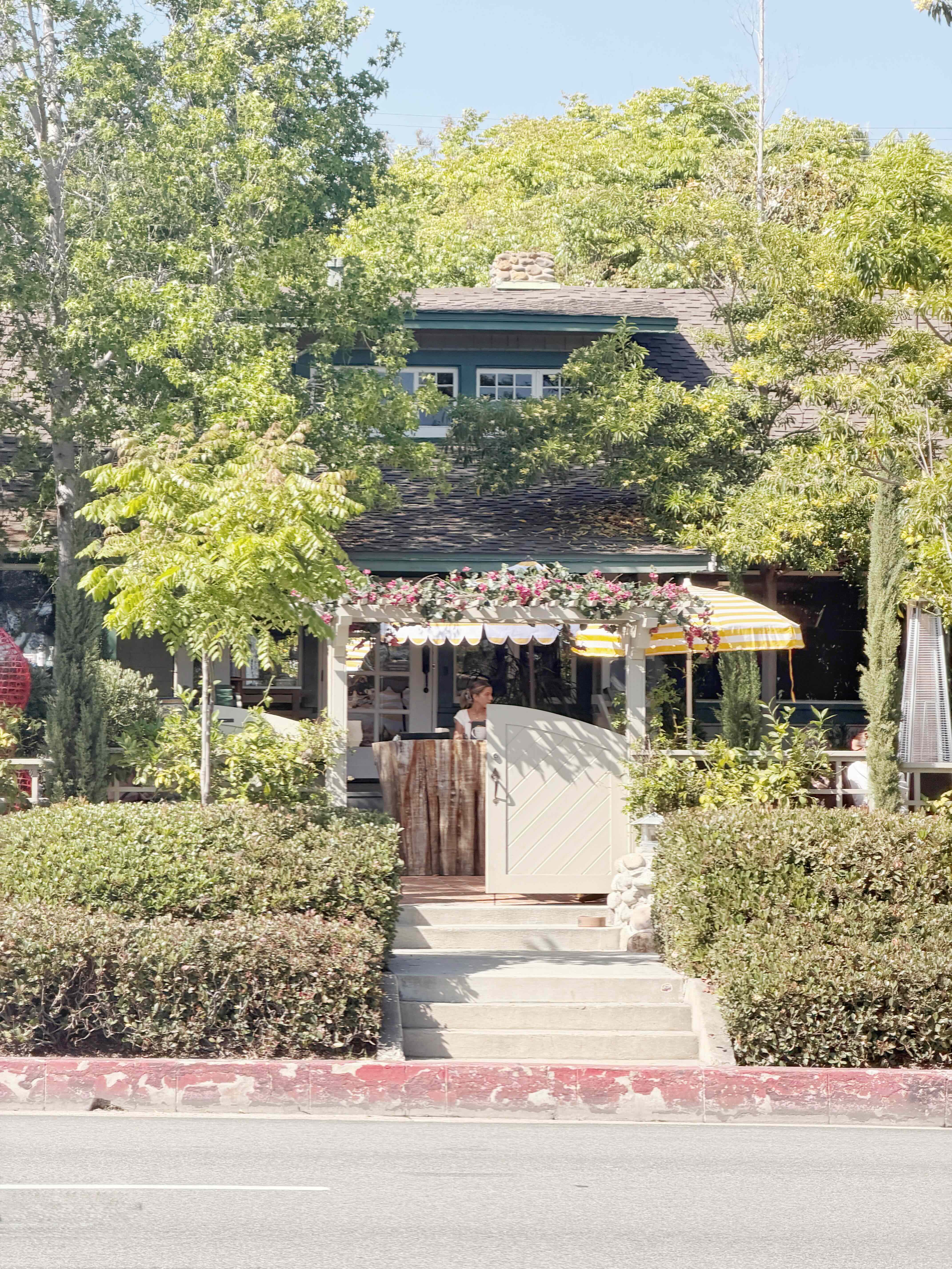 Truly Pizza Laguna Beach exterior on North Coast Highway — white craftsman cottage with green roof, scalloped yellow-and-white patio umbrella, picket fence and flower-lined patio across from the Laguna Art Museum