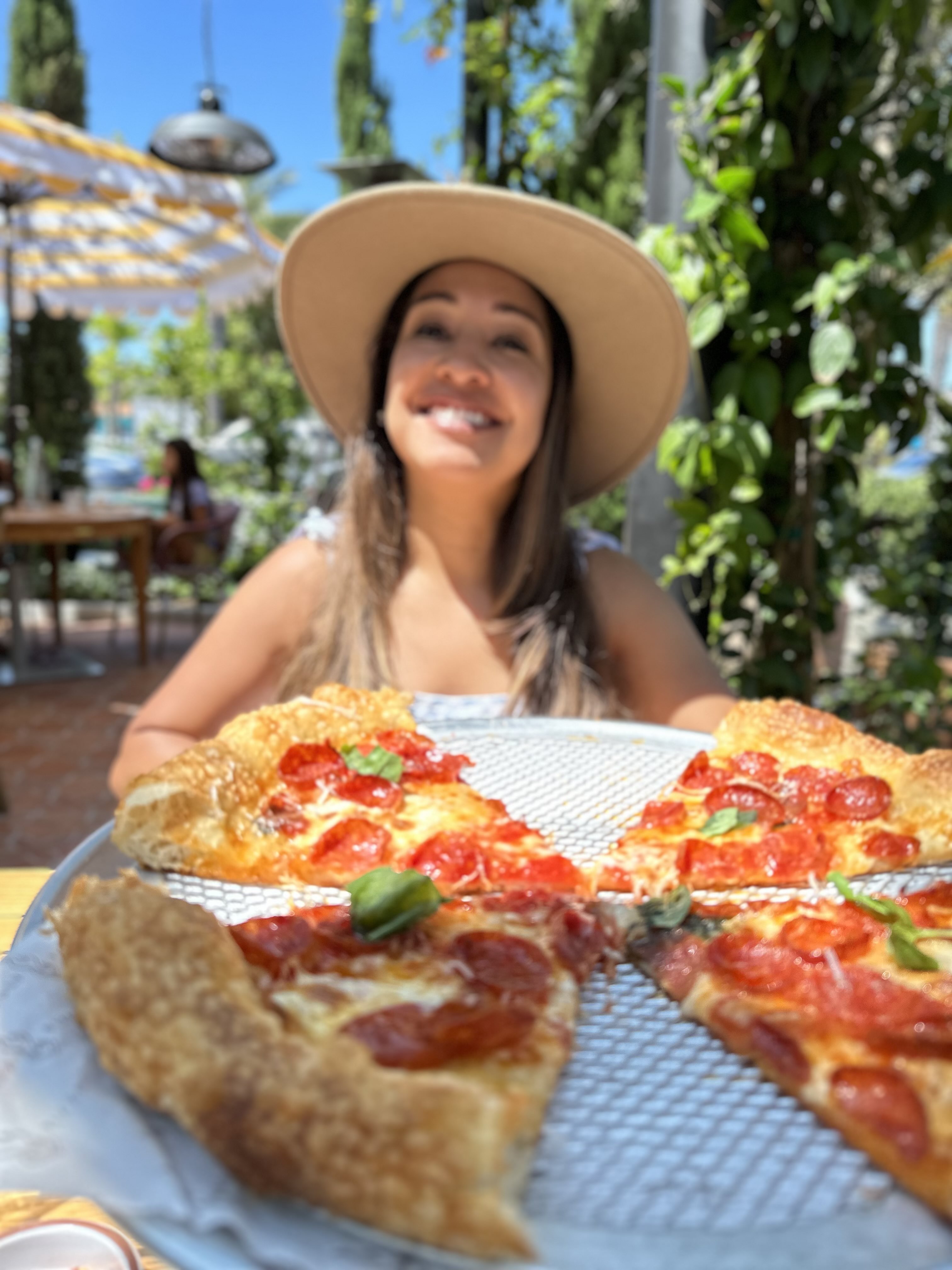A diner smiling behind a large pepperoni pizza on a metal serving rack at Truly Pizza Dana Point — yellow-striped patio umbrella and lush greenery in the background