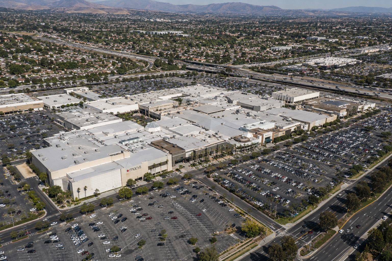 Aerial view of Brea Mall in Brea, Orange County: mall structure flanked by extensive parking, surrounding residential neighborhoods, and the San Gabriel foothills in the background.