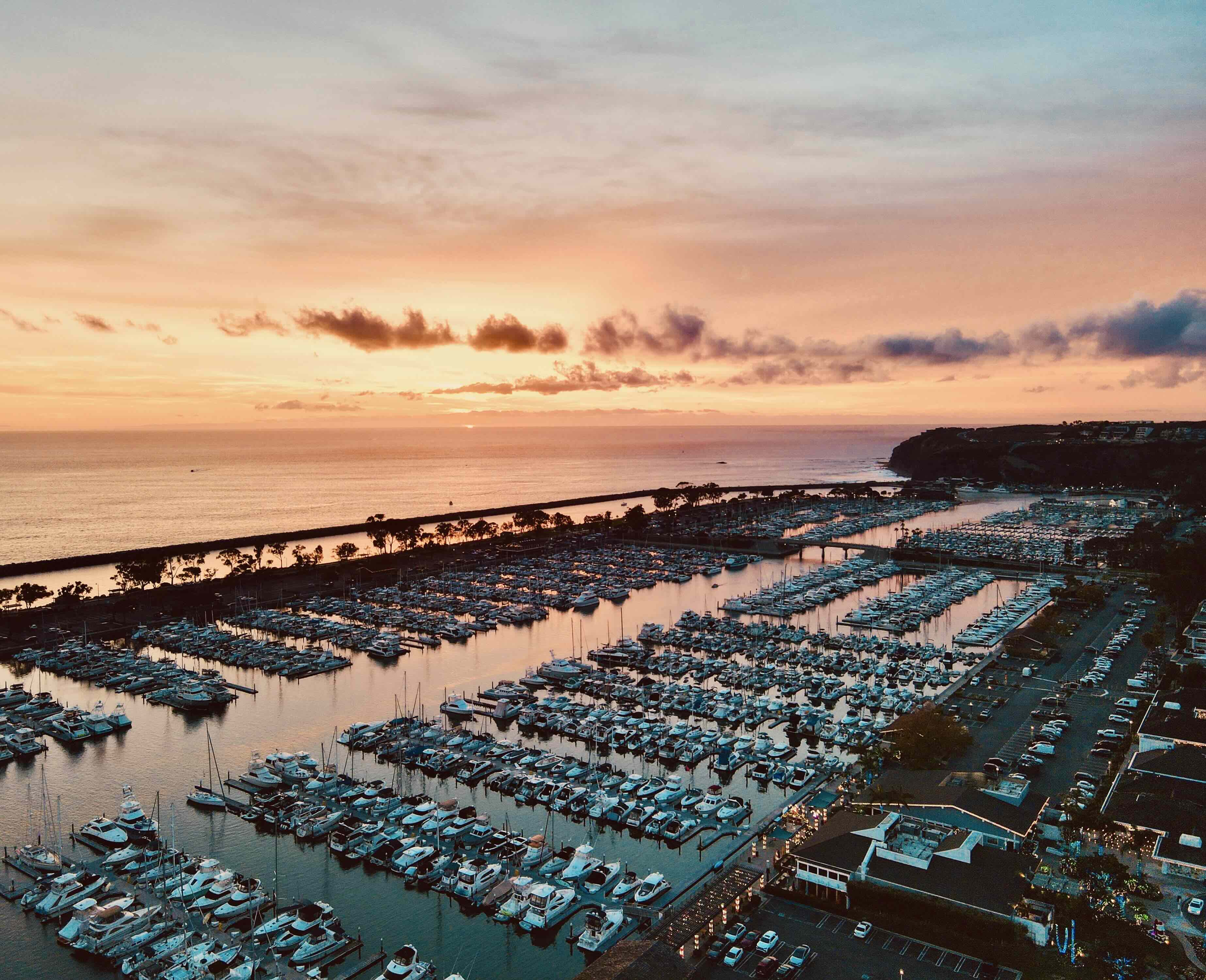 Dana Point Harbor at sunset: aerial view of hundreds of moored yachts and sailboats in the marina, the protective breakwater jetty, the Pacific Ocean beyond, and coastal bluffs framing the east side of the harbor.