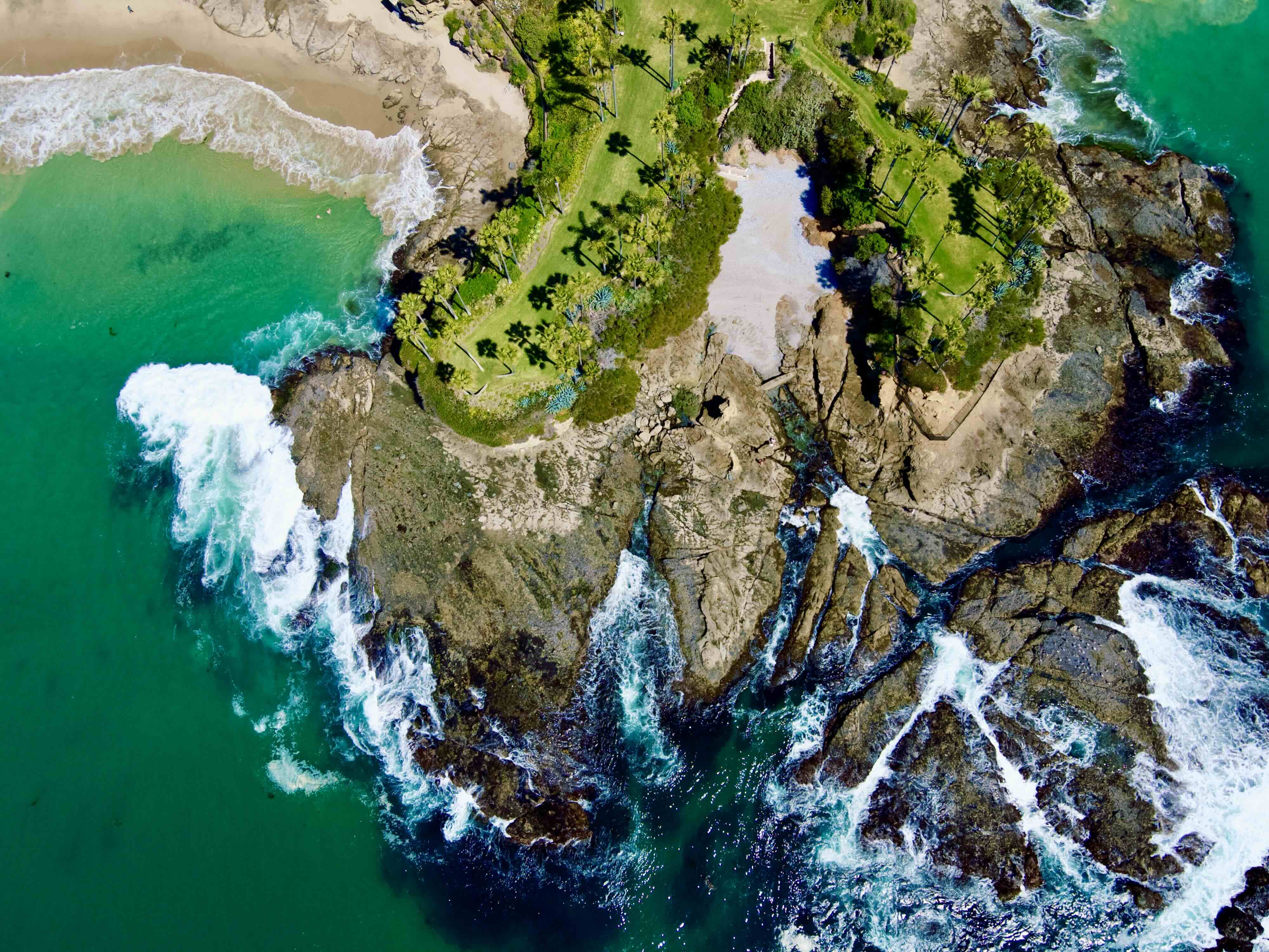 Aerial view of Laguna Beach's coastline: a grass-and-palm-topped headland projecting into the Pacific, dramatic rocky cliffs and tide pools, turquoise water with surf breaking on the rocks, and a hidden cove beach to the upper left.