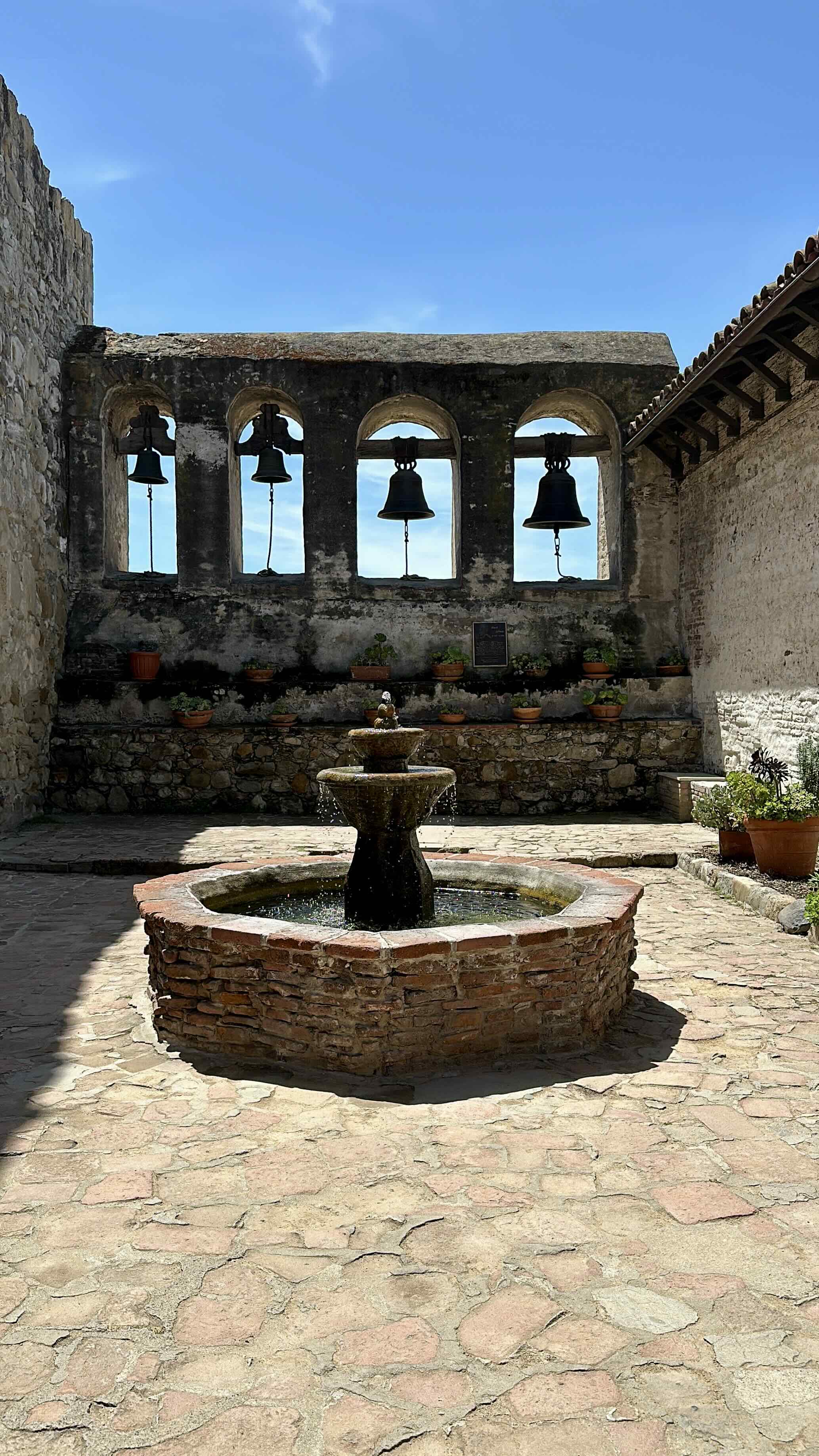 The historic four-bell campanario at Mission San Juan Capistrano rising above a flagstone courtyard, with a two-tiered stone fountain at center and potted succulents lining the weathered stone wall under a clear blue sky.