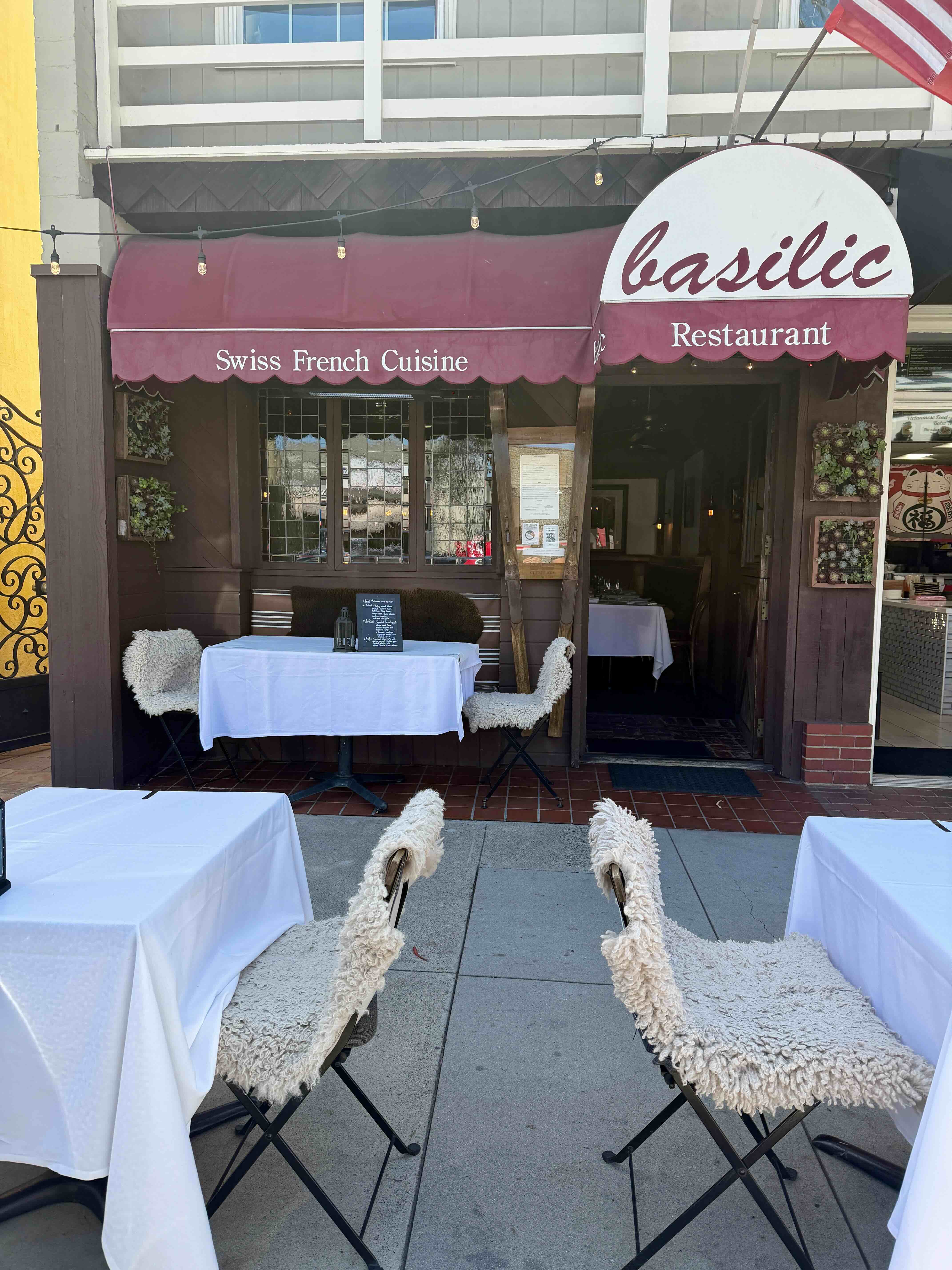 Basilic Restaurant on Balboa Island — burgundy awning over the Swiss-French dining storefront on Marine Avenue with sidewalk tables, fluffy white chair covers, and the American flag flying above