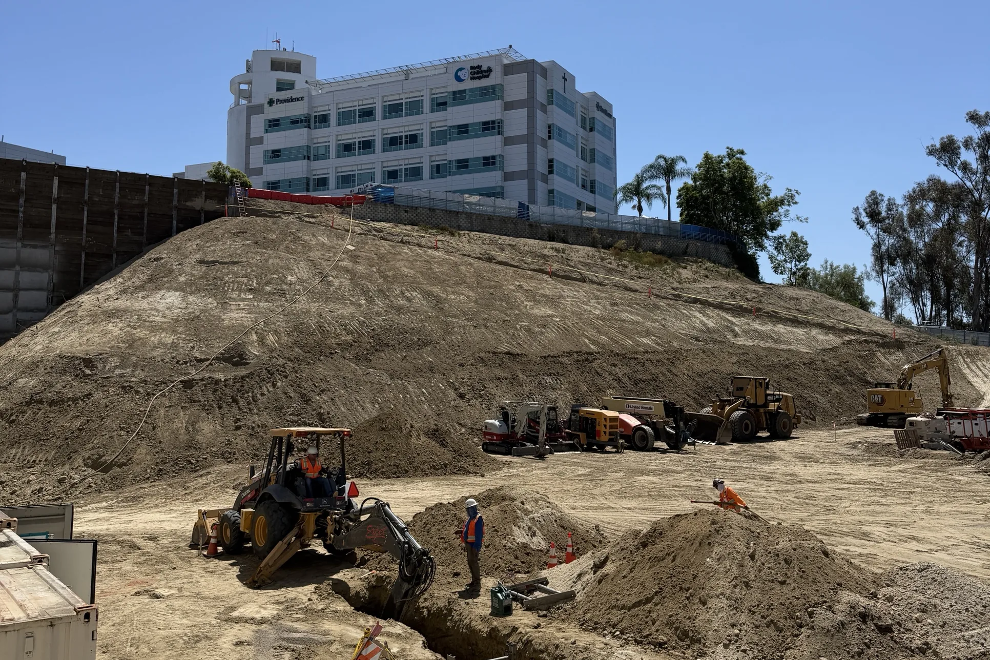 A backhoe digs a trench on a construction site as workers, an excavator and a loader work the graded dirt below a Providence Mission Hospital building with Providence and Rady Children's Hospital signage in Mission Viejo.