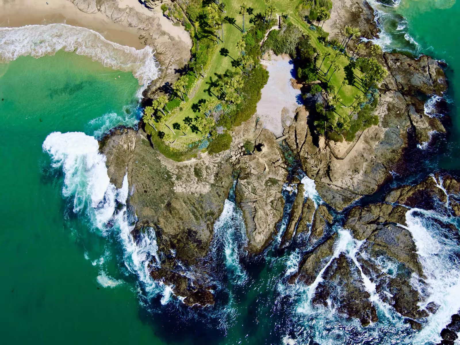 Aerial view of Laguna Beach's coastline: a grass-and-palm-topped headland projecting into the Pacific, dramatic rocky cliffs and tide pools, turquoise water with surf breaking on the rocks, and a hidden cove beach to the upper left.