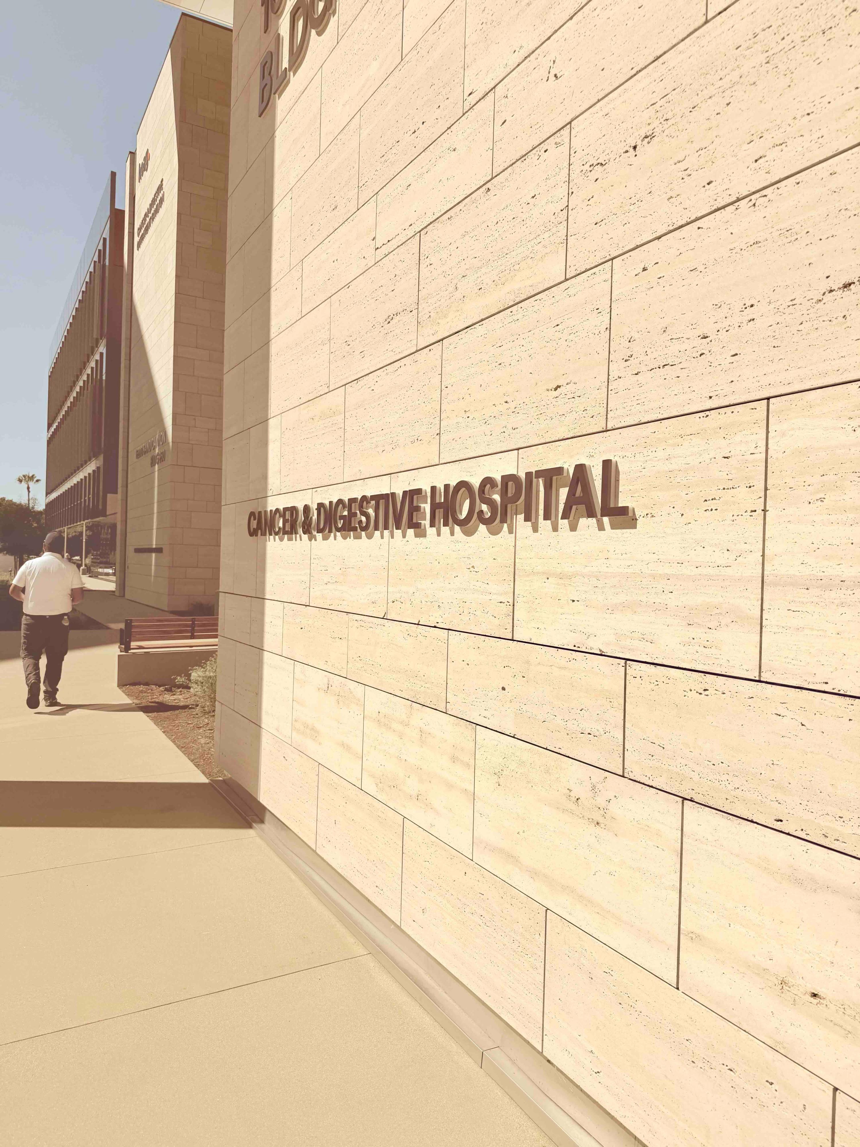 Cancer & Digestive Hospital lettering on the travertine wall of the new pavilion at Hoag Hospital Irvine, with a pedestrian walking along the sidewalk.