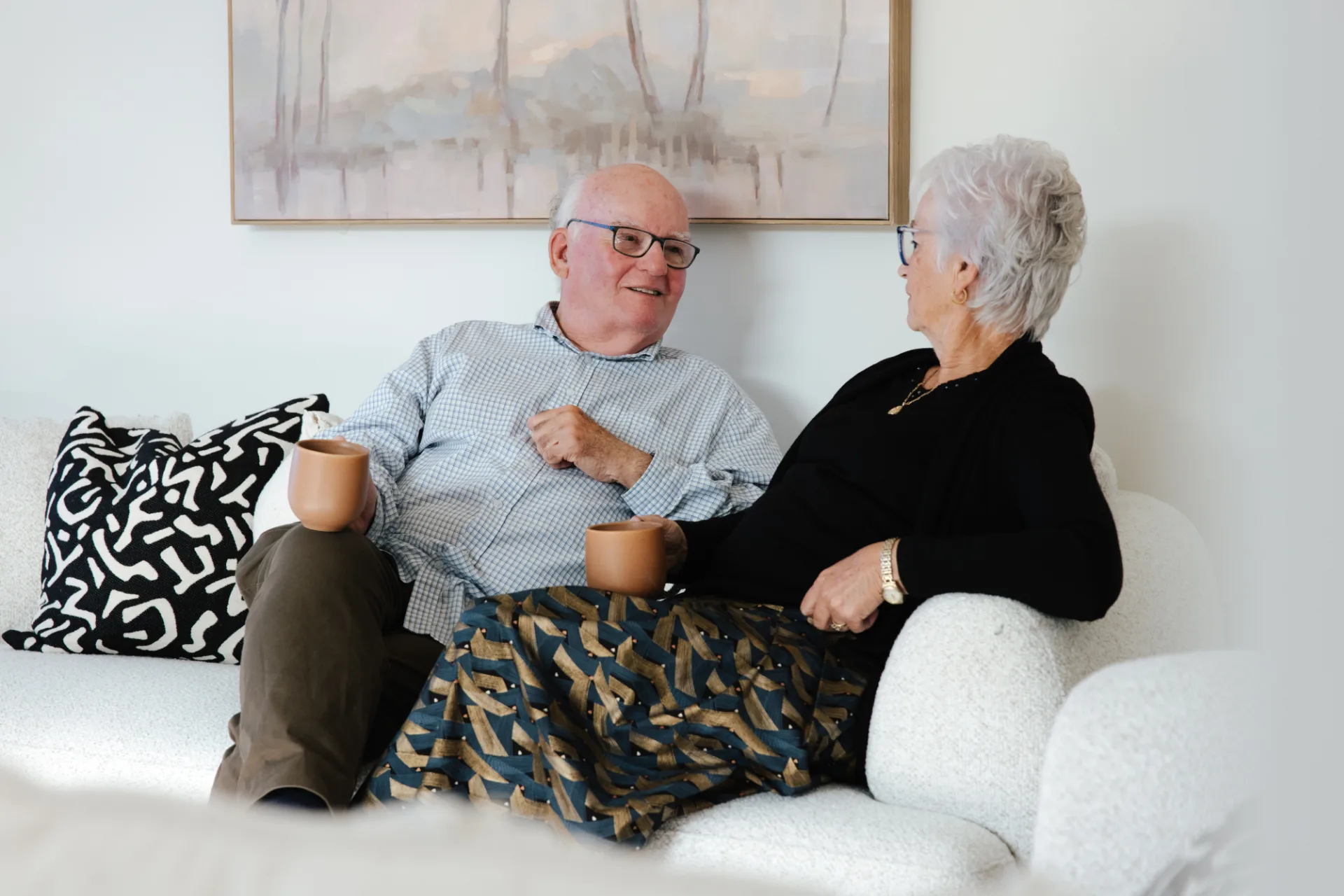 Elderly man and woman sitting on a white couch, holding coffee mugs and chatting.