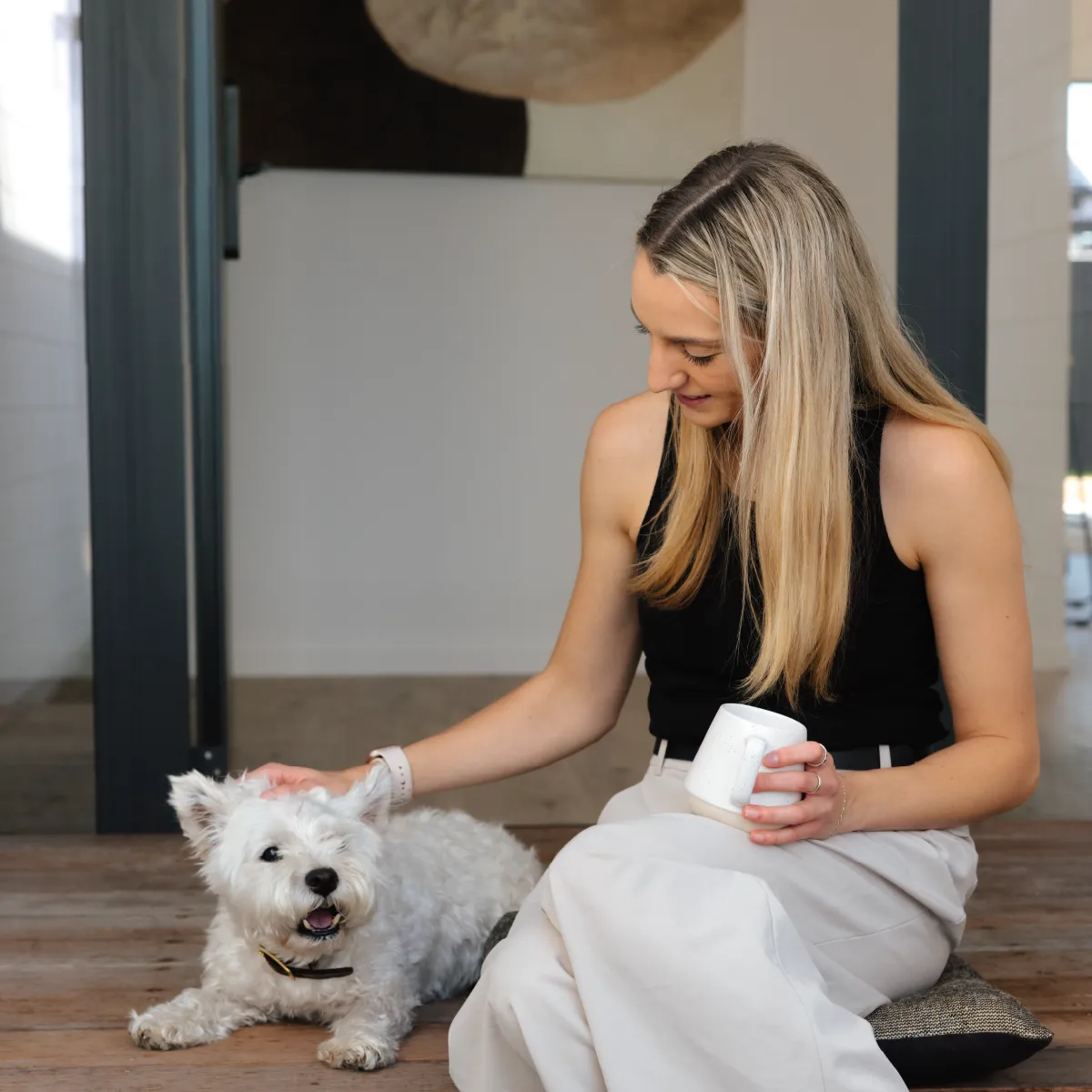 Woman in black top sitting on cushion petting a happy white dog while holding a white mug.