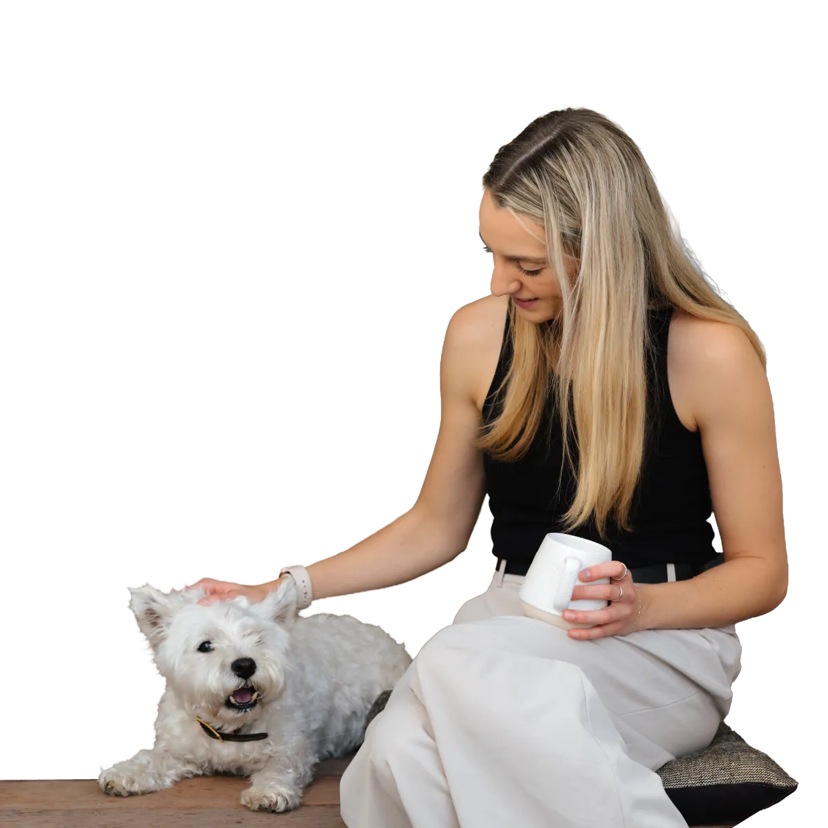 Smiling woman in black top petting a white dog while holding a white mug, sitting on a cushion.