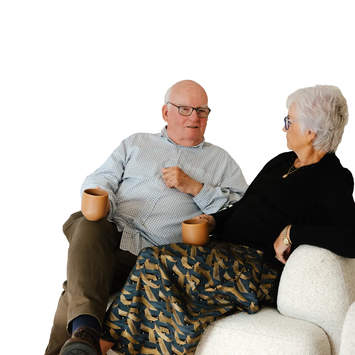 Elderly man and woman sitting on a couch holding mugs, engaged in conversation.