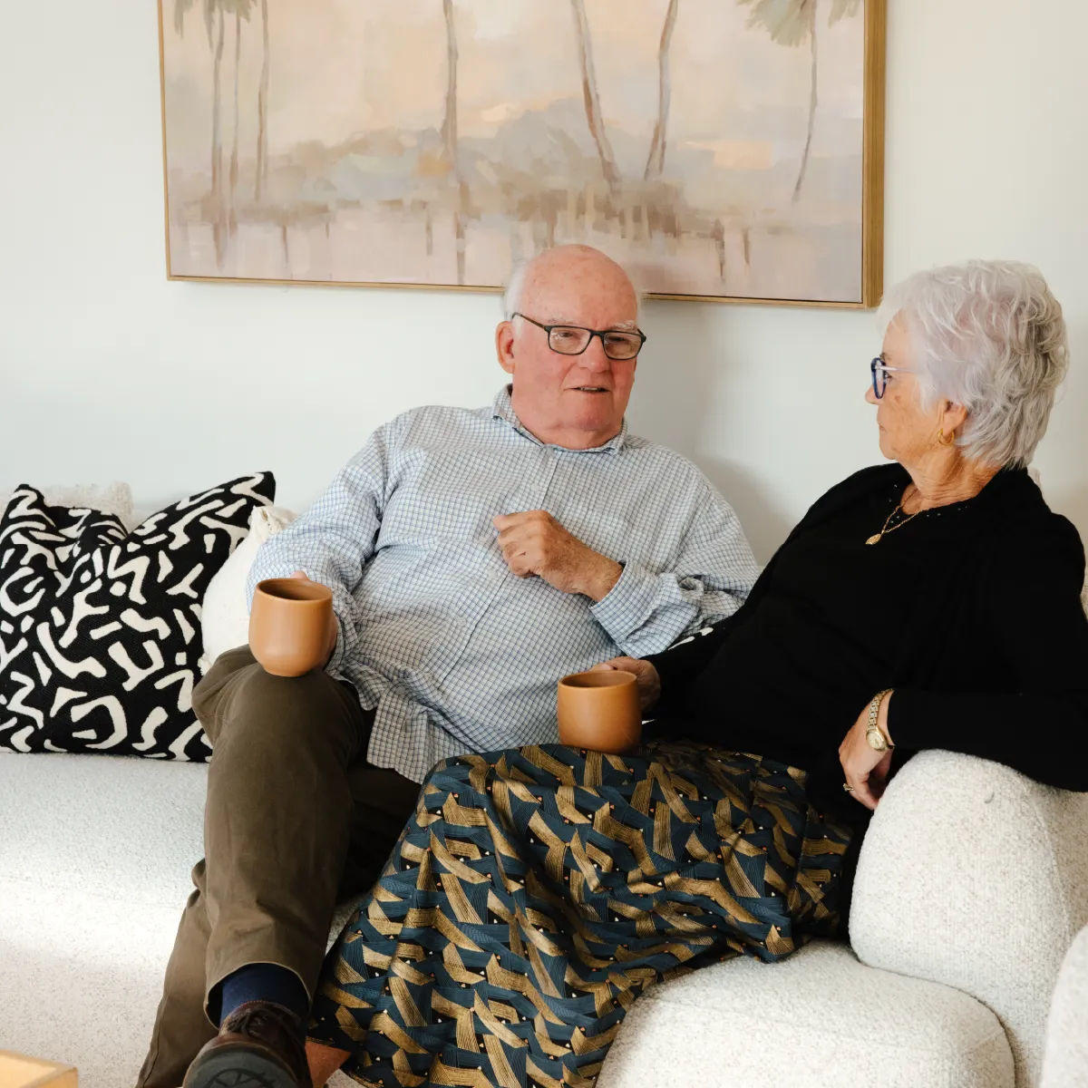 Elderly man and woman sitting on a couch, each holding a brown mug, engaged in conversation.
