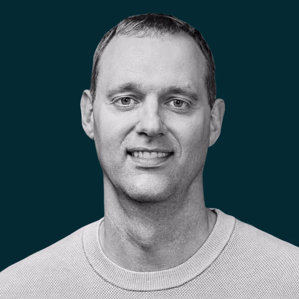 Black and white headshot of a smiling man with short hair wearing a crew neck sweater against a dark background.