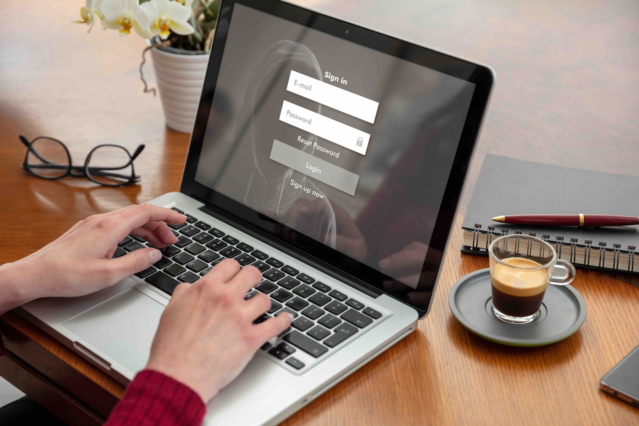 Person typing on a laptop keyboard with a sign-in screen displayed, a cup of espresso, glasses, and notebook on a wooden table.