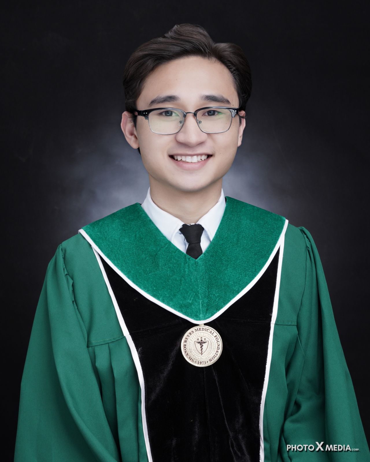 Smiling graduate wearing glasses and a green and black academic gown with a medical foundation emblem.