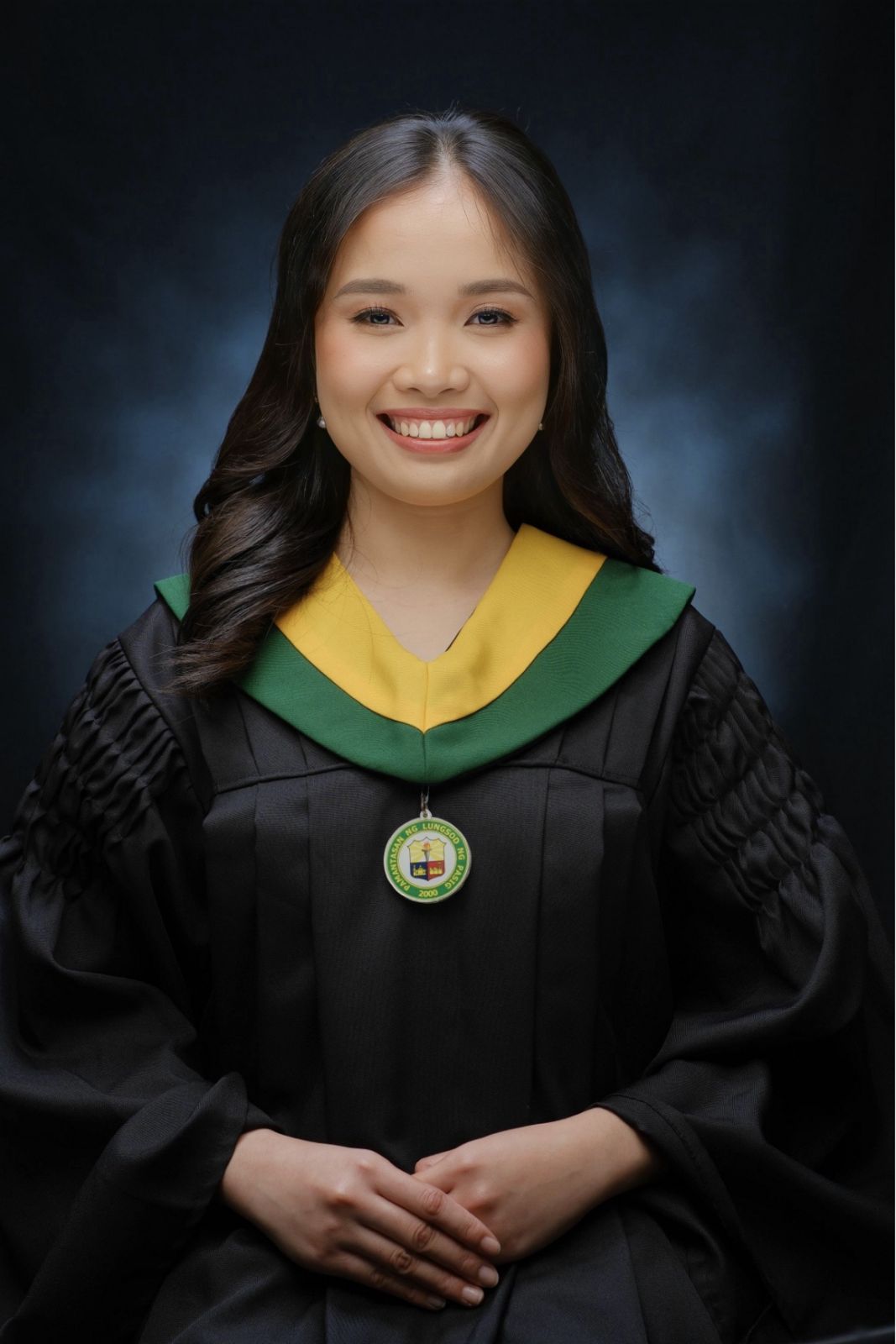 Smiling young woman in a black graduation gown with green and yellow collar against dark background.