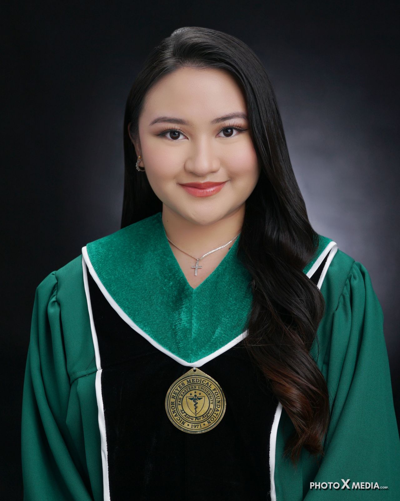 Smiling woman in green and black graduation gown with a cross necklace on black background.